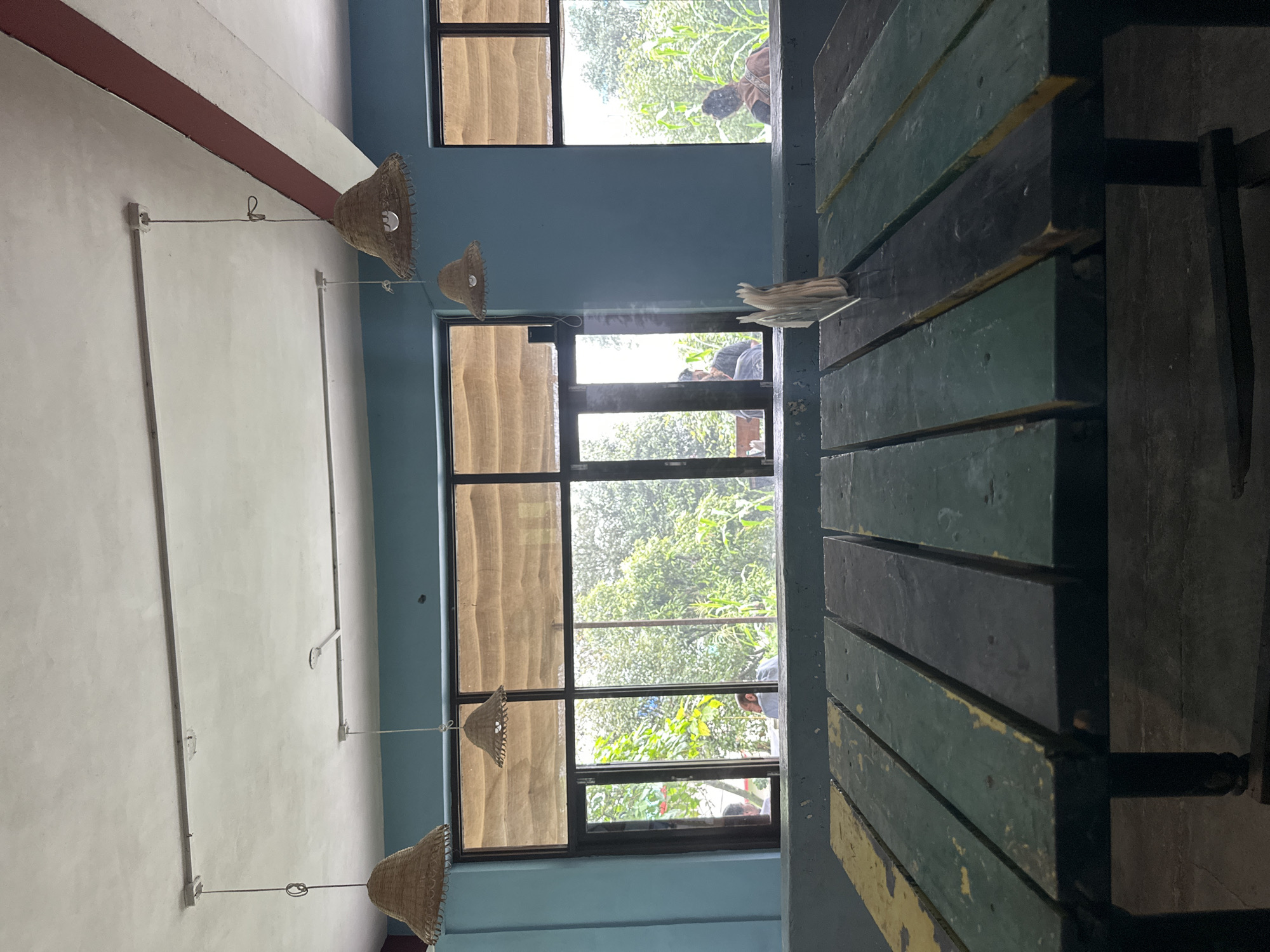 The image depicts the interior of a restaurant with a view of greenery through large windows. In the foreground, a weathered wooden table with planks painted in alternating shades of dark green, black, and yellow is visible. A napkin holder stands on the table. Above the table, the blue walls are adorned with three woven straw pendant lamps hanging from the ceiling. The windows, divided into multiple panes, reveal lush trees and foliage outside, with some glimpses of individuals in the distance. One window has a glass door that appears to open to the outside. The ceiling is white with a reddish-brown band along the edge. The overall impression is a casual, perhaps rustic, dining environment.