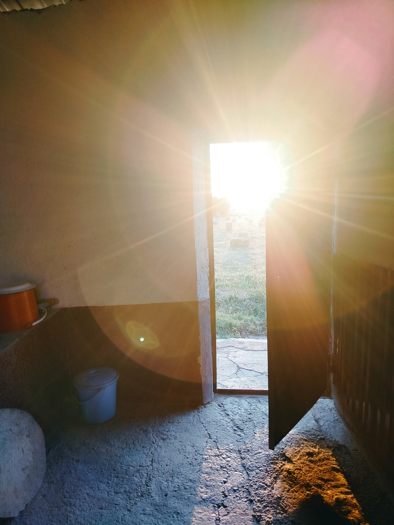 The image depicts an interior view looking out through an open doorway. Intense sunlight streams in from the doorway, creating lens flares and casting bright rays across the scene. The interior appears to be a simple, possibly rustic, room with rough plaster walls. On the left side of the room, there is an orange bucket with a white lid placed on a shelf or ledge, and a white bucket on the floor. A large, round, grey stone is also visible on the floor to the left. The floor itself is a rough, concrete surface. Through the doorway, a grassy area with some scattered rocks is visible, illuminated by the bright sunlight. The scene is set in an unspecified location within the city of Көлкент, Kazakhstan, though no specific landmarks are identifiable from the image. There are no people or animals visible in the frame.