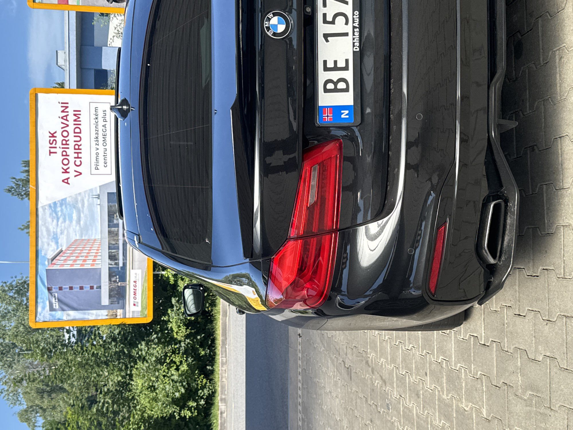 This image shows the rear passenger side of a black BMW sedan parked on a paved surface, likely a parking lot, with a billboard in the background. The billboard features an advertisement with text in Czech, mentioning "TISK A KOPÍROVÁNÍ V CHRUDIMI" which translates to "PRINTING AND COPYING IN CHRUDIM". The billboard also displays the words "Přímo v zákaznickém centru OMEGA plus", meaning "Directly at the OMEGA plus customer center."
The car has a Norwegian license plate that reads "BE 157" and the text "Dahles Auto" beneath it. The BMW emblem is visible on the trunk. The environment appears to be outdoors, with greenery and trees visible behind the billboard and a clear blue sky overhead. The pavement is made of interlocking concrete blocks. The overall scene is that of a vehicle parked near an advertisement in the city of Chrudim, Czechia.