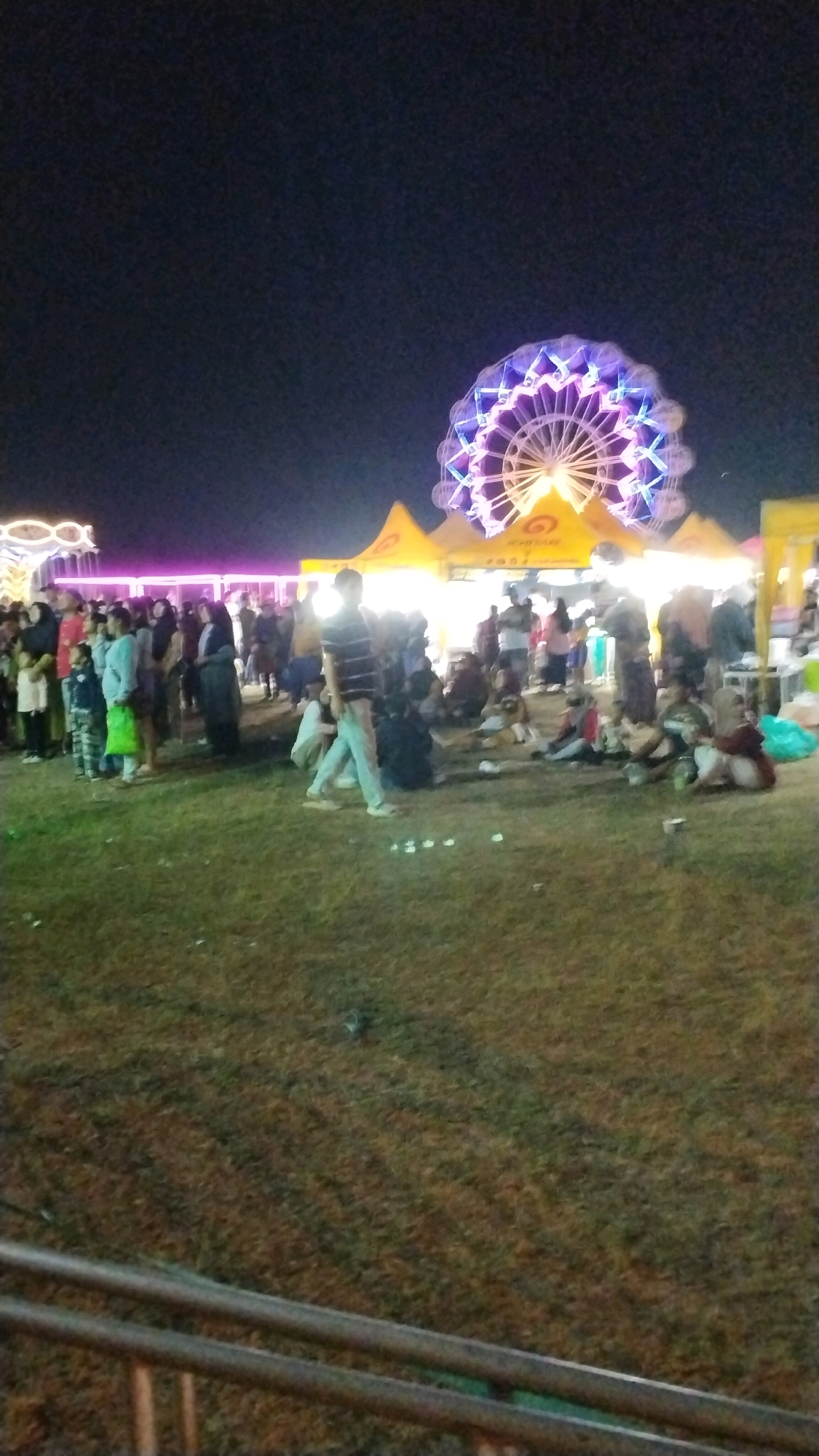 The image shows a night scene at an outdoor event, possibly a fair or festival, in Canggu, Indonesia. A large Ferris wheel, illuminated with purple and white lights, is visible in the background. In the midground, several yellow tents with logos are set up. A crowd of people, of various ages and genders, are gathered in the scene. Some are standing and walking, while others are sitting on the grass. The ground is covered in grass, and there is a metal railing in the foreground. The lighting is dim, with the main illumination coming from the Ferris wheel and the tents.