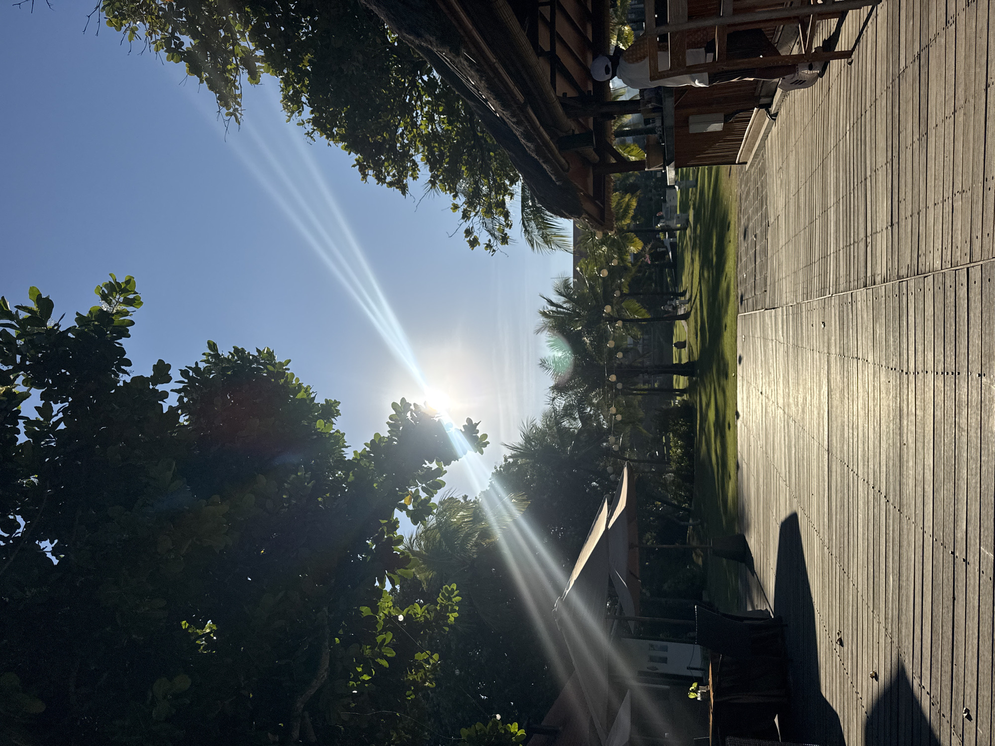 The image captures a sunlit outdoor scene, likely in Kuta Selatan, Indonesia, given the presence of tropical vegetation. The sun, positioned near the center of the frame, emits strong rays that are diffused by trees. To the left of the sun, lush green trees dominate. A wooden deck occupies the right side of the image, with visible nailheads and shadows cast upon it. In the middle ground, palm trees line a grassy area. A building with a thatched roof and open structure is situated to the right of the palm trees, containing figures who appear to be staff or patrons, partly obscured. The sky is a clear blue.