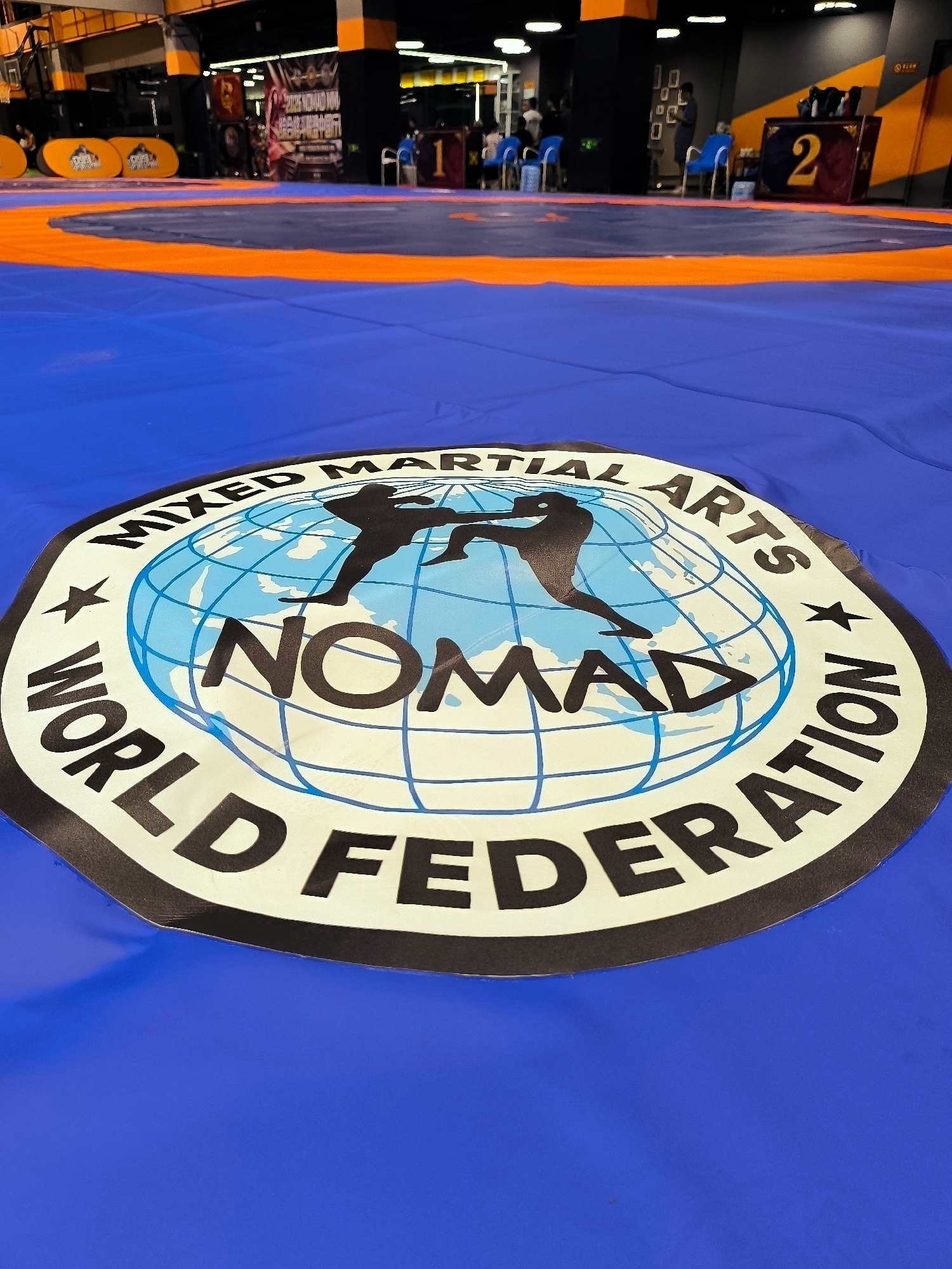 The image shows a close-up of a blue wrestling mat with a circular logo in the center. The logo features a globe with two silhouetted figures engaged in a martial arts pose. Text around the globe reads "MIXED MARTIAL ARTS WORLD FEDERATION NOMAD." The mat is set up in what appears to be an indoor arena or gymnasium, with orange and blue boundaries visible in the background. Spectator seating and event signage are also present in the blurred background. The lighting suggests an indoor, possibly artificial, environment.