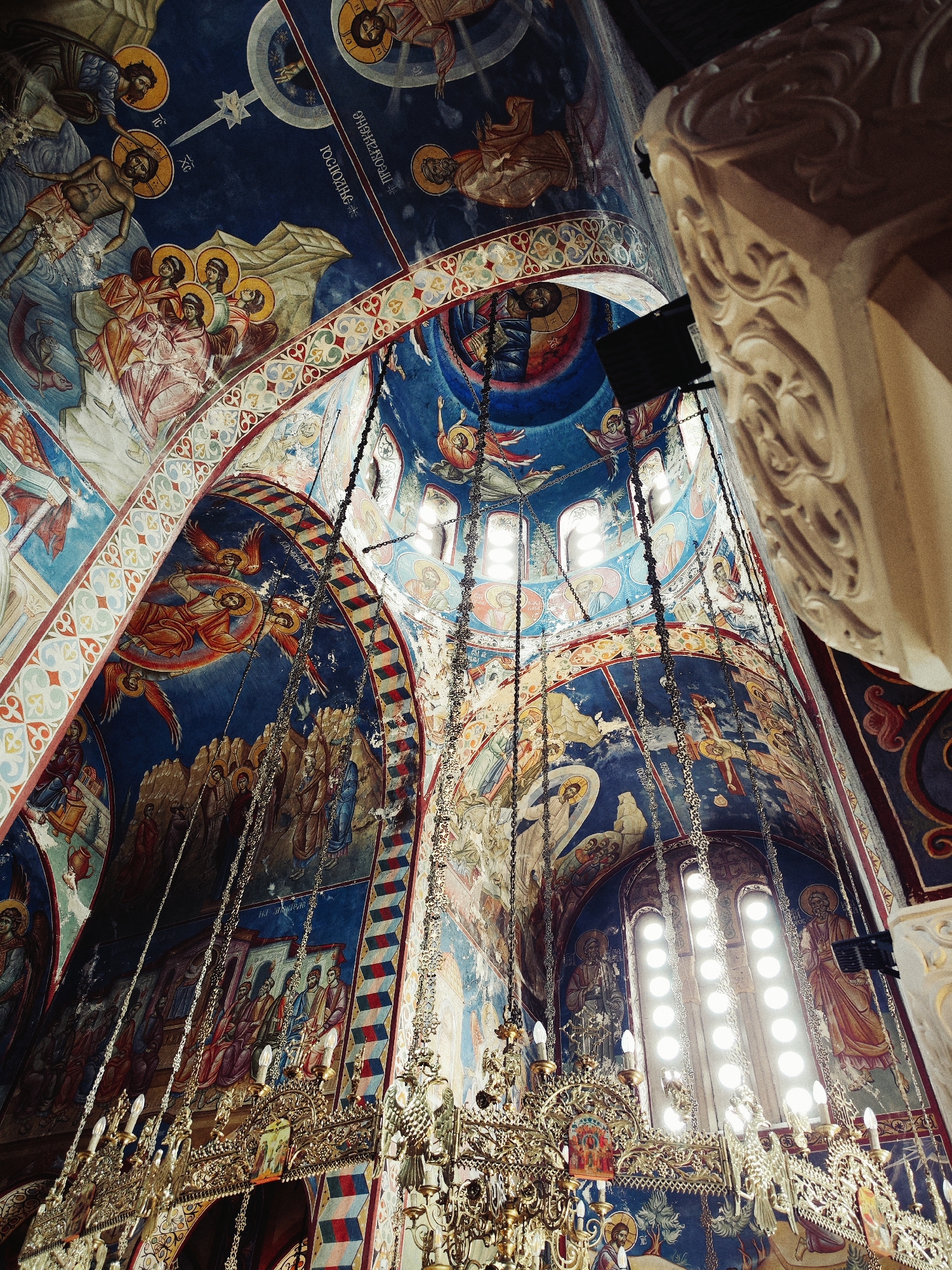 The image showcases the interior of the Hercegovačka Gračanica monastery in Trebinje, Bosnia and Herzegovina. The camera is positioned at a low angle, capturing the intricate ceiling frescoes, which depict biblical scenes and religious figures in vivid blue, red, and gold tones. The architecture includes arches decorated with geometric patterns. Large chandeliers with chains and ornate metalwork hang from the ceiling. A section of a carved stone pillar is visible in the foreground on the right. Natural light streams in through a row of arched windows.