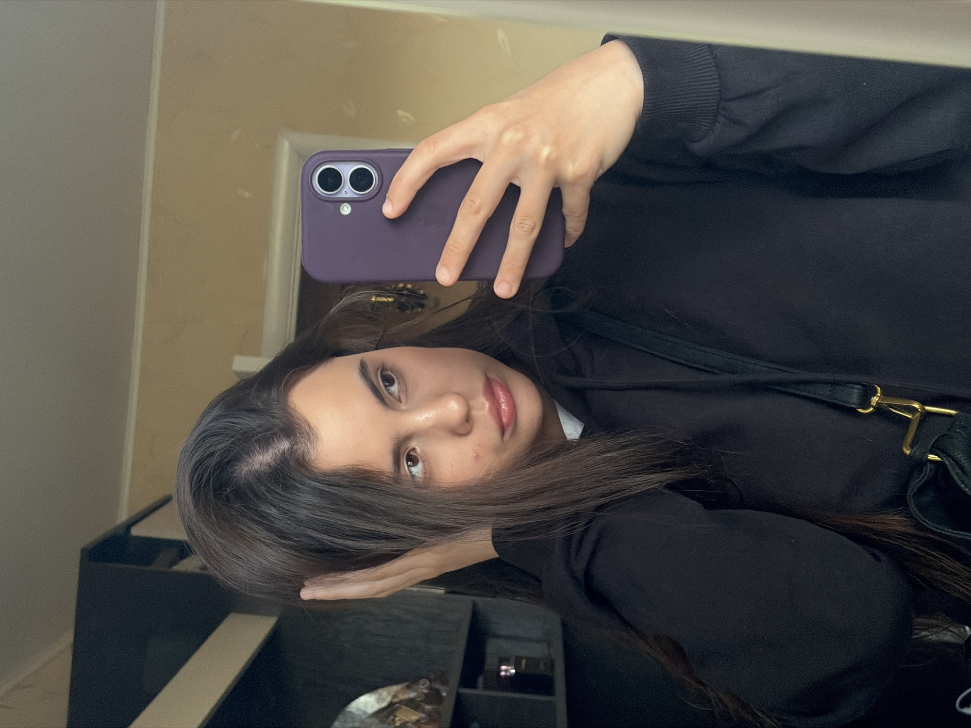 A mirror selfie captures a young woman from the chest up, rotated 90 degrees counter-clockwise. She has long, dark brown hair, parted on the side, that flows over her shoulder and partially covers her face. Her eyes are dark brown, and she has a calm, neutral expression with slightly parted lips. She appears to be looking directly at her reflection in the mirror.
She is wearing a black, long-sleeved top, possibly a hoodie, with a ribbed cuff visible on her right arm. A black shoulder bag with a prominent gold metal clasp is visible on her left shoulder. She holds a purple smartphone, likely an iPhone with two distinct camera lenses and a flash, in her right hand, which is raised to take the photograph.
The background, a reflection in the mirror, shows an indoor setting with light beige walls and dark furniture, possibly a dresser or cabinet. Some indistinct objects, including what appears to be a silver tray and small containers, are visible on the furniture. There is no visible text in the image.