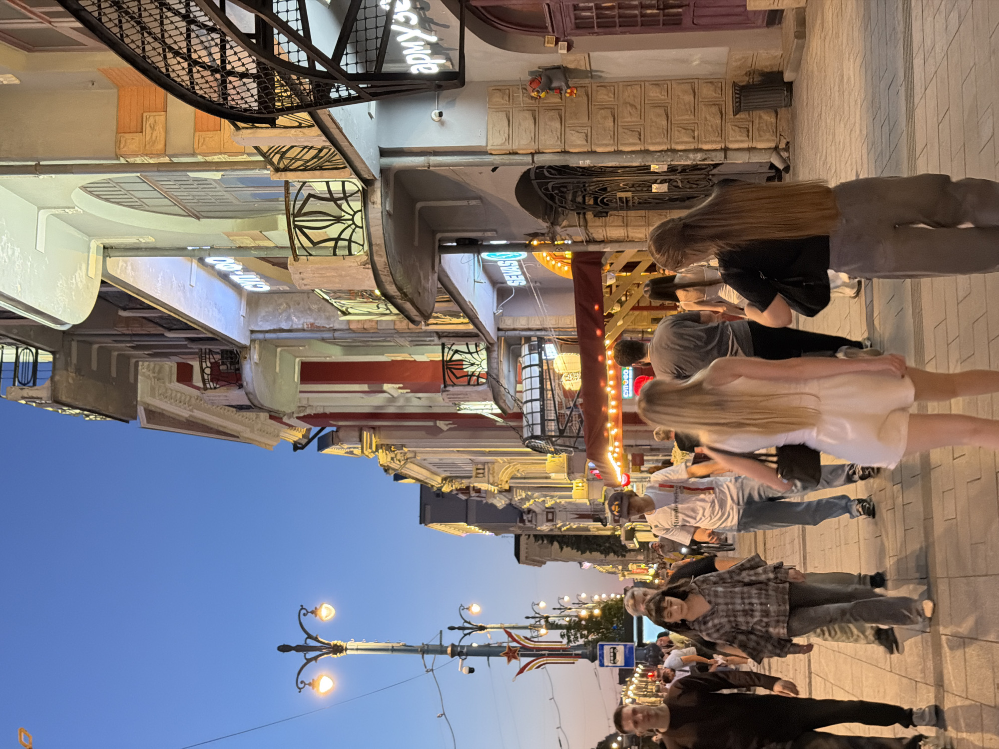 The image depicts a street scene in Novaya Usman', Russia, at dusk. The sky is a clear, deep blue, transitioning to a lighter hue near the horizon. Ornate streetlights with glowing bulbs illuminate the paved walkway and the facades of buildings on either side. The buildings showcase a mix of architectural styles, with some featuring classical elements like columns and decorative molding, while others have more modern designs with large windows and neon signs.
Several people are visible, walking along the street. Some are in the foreground, while others are further back, blending into the crowd. The people appear to be engaged in casual activity, likely enjoying an evening stroll or socializing. Their attire suggests a comfortable, perhaps warm, evening.
One building has a prominent neon sign that is partially legible, appearing to spell out "СИЯНЬЕ" (Siyanye, meaning "radiance" or "shine" in Russian). Another sign reads "арм.узе" (arm.uze), possibly an abbreviation or part of a longer word. The overall atmosphere is lively and inviting, with the warm glow of the lights and the presence of people suggesting a pleasant urban environment. The time of day is clearly evening, as indicated by the twilight sky and the illuminated streetlights.