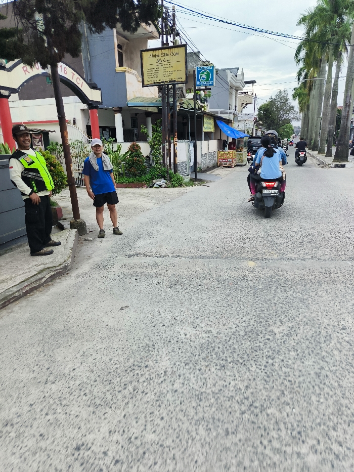 The image shows a street scene in Kecamatan Dayeuhkolot, Indonesia (latitude: -6.963, longitude: 107.596). A man in a black uniform and a yellow vest stands on the left side of the street, looking directly at the camera. He has a walkie-talkie on his belt. Another man wearing a blue shirt and a white hat is standing on the sidewalk to the right of the image, looking towards the street. Behind them, a building with a sign that reads "Aisha Skin Care Salon" is visible. A motorbike with two passengers is driving towards the camera on the street. In the distance, there are more buildings and trees along the street. The sky is cloudy.