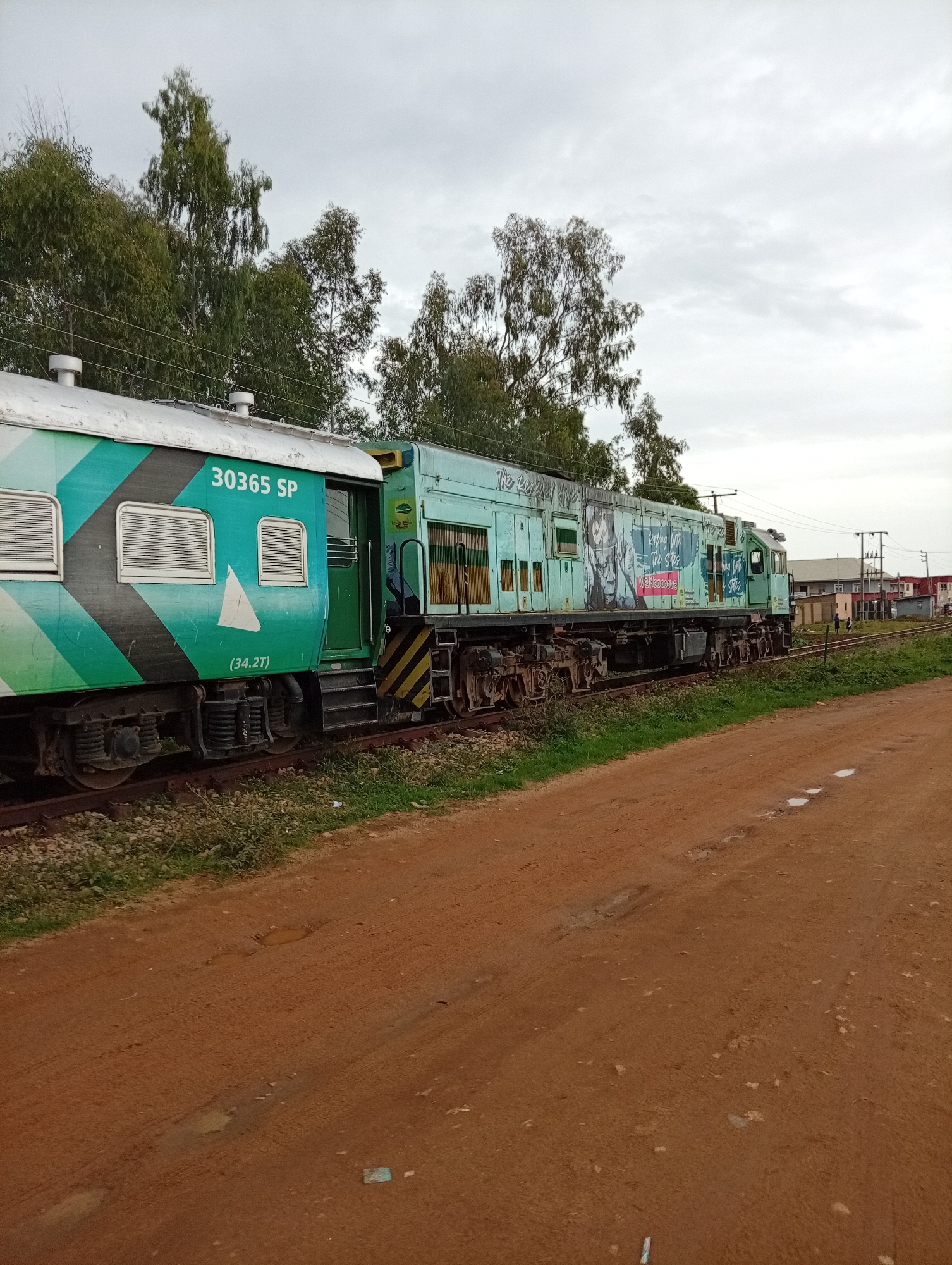 An abandoned train sits on tracks in Gura Top, Nigeria, under a cloudy sky. The train is light blue and features various graphics and text, including "30365 SP" and "(34.2T)". A green carriage is attached to the front. The train is surrounded by overgrown grass and dirt roads, with trees and some buildings visible in the background. The scene appears to be a railway yard or a disused section of track.