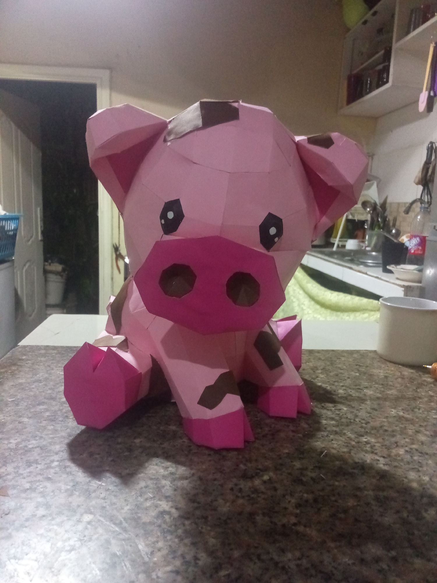 A close-up shot shows a papercraft pink pig on a speckled countertop. The pig has brown spots and a darker pink snout. Its features are cartoonish, with large black eyes and triangular ears. The setting appears to be a kitchen, with a partially visible doorway leading to an outdoor area in the background to the left. Behind the pig, there is a sink, dishes, utensils hanging, and shelves with jars and other kitchen items. A white mug is on the counter to the right. The location cannot be ascertained.