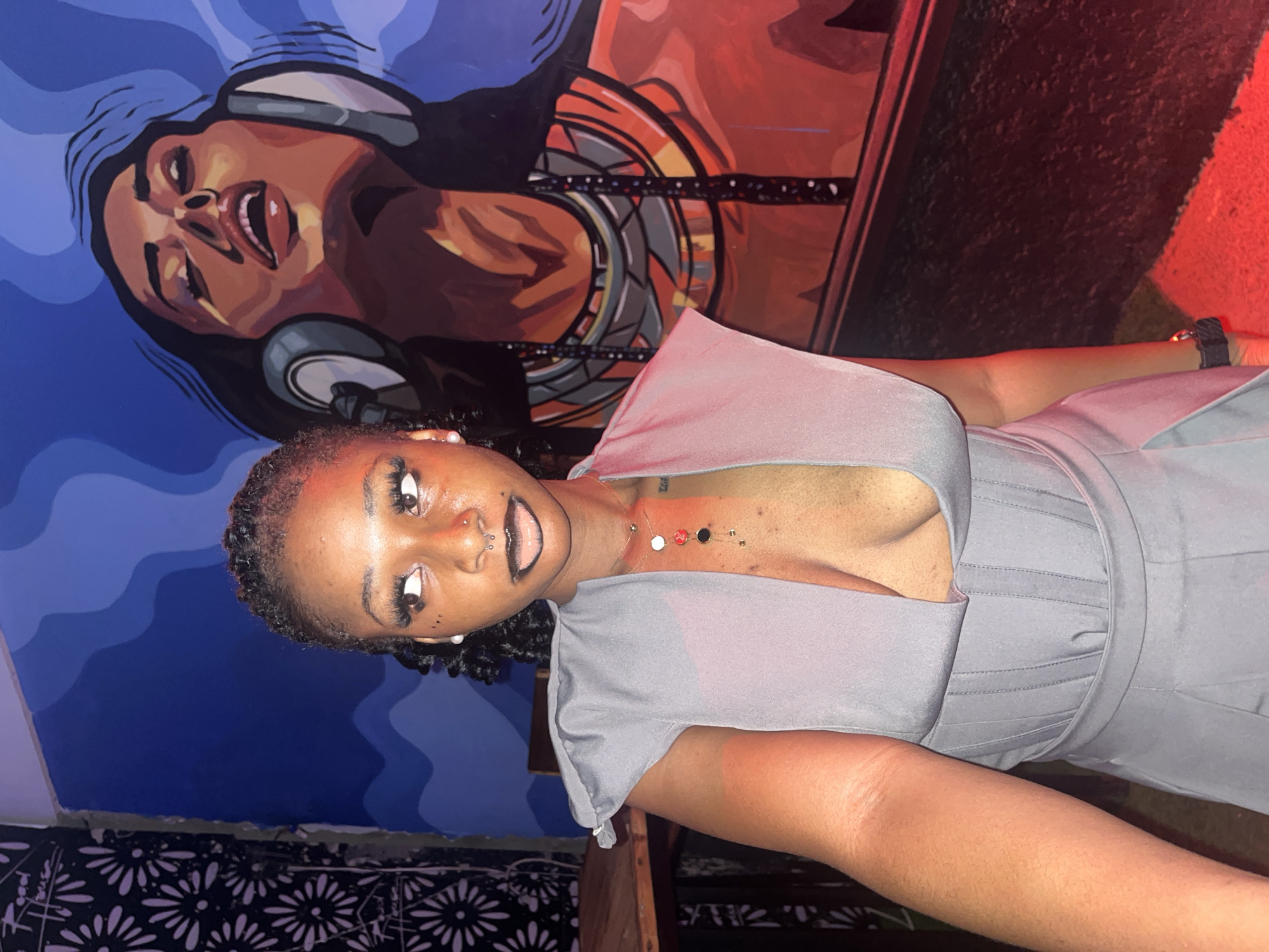 A young Black woman with braided hair, wearing a grey v-neck dress and dark lipstick, poses in front of a mural. The mural depicts a woman with headphones and elaborate jewelry. The background features a wall with a floral pattern on the left and a blue wavy design on the right. A table and chairs are partially visible on the far left. Red lighting illuminates the lower portion of the scene. The image appears to be taken indoors, possibly in a bar or nightclub, in the city of Howayi, Nigeria.