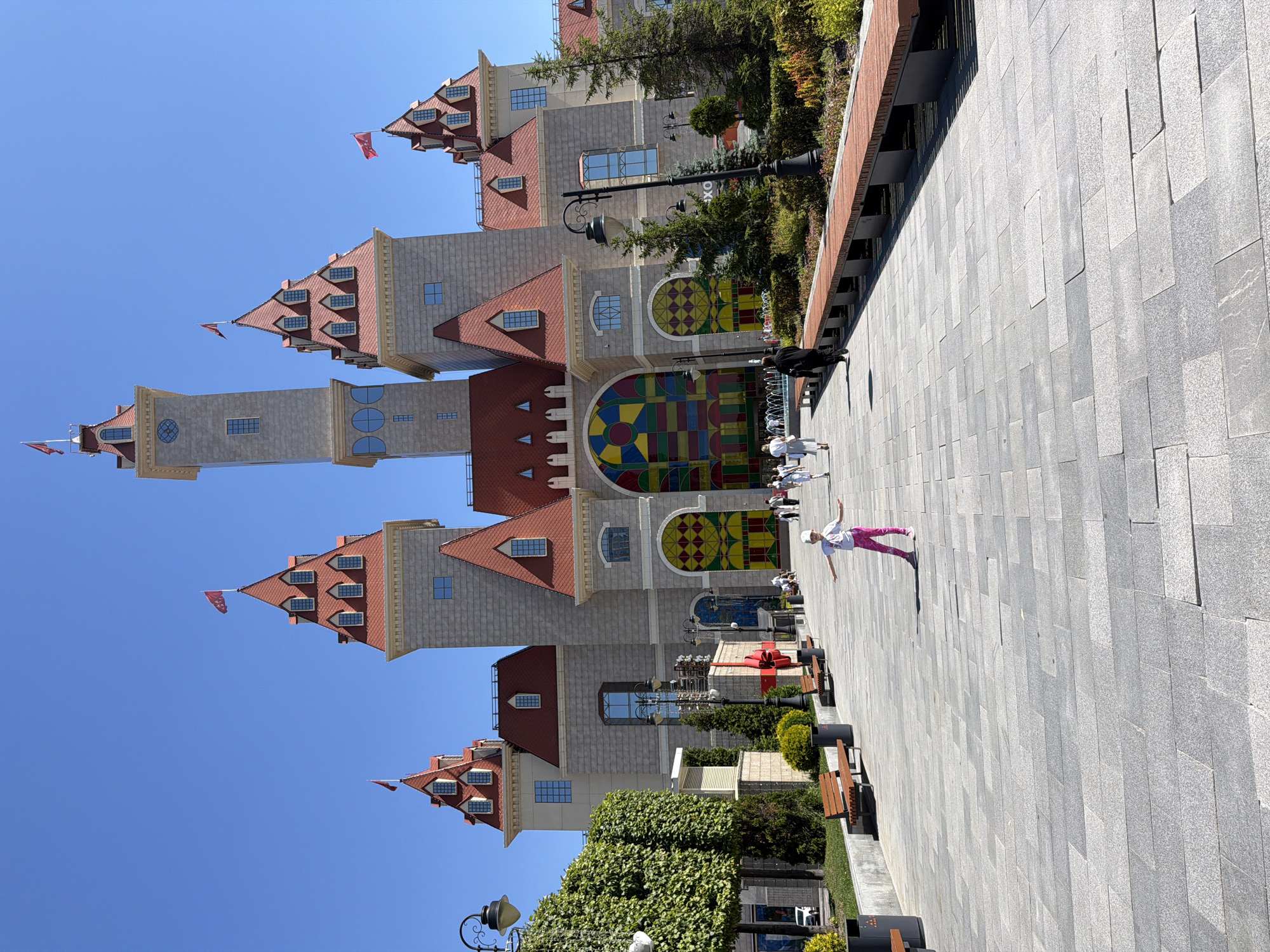 The image depicts a bright, sunny day in front of a whimsical, castle-like building. The architecture features multiple towers with red roofs, dormer windows, and flags flying from the highest points. A prominent feature is a large, arched entrance filled with colorful stained glass. In the foreground, a young girl in pink pants and a white shirt is posing with outstretched arms on a paved plaza. Several other people are walking in the middle ground, and park benches and greenery line the path. The environment suggests a public space, possibly a park or an entertainment venue, designed to evoke a fairy tale aesthetic. The building's design is reminiscent of amusement park architecture, and its scale suggests it might be a significant landmark. There are street lamps and decorative elements that contribute to the festive atmosphere. The overall impression is one of a vibrant and engaging public space, likely designed for families and entertainment. Based on the architectural style and public space design, this location is likely Moscow, Russia, possibly a theme park or entertainment complex.