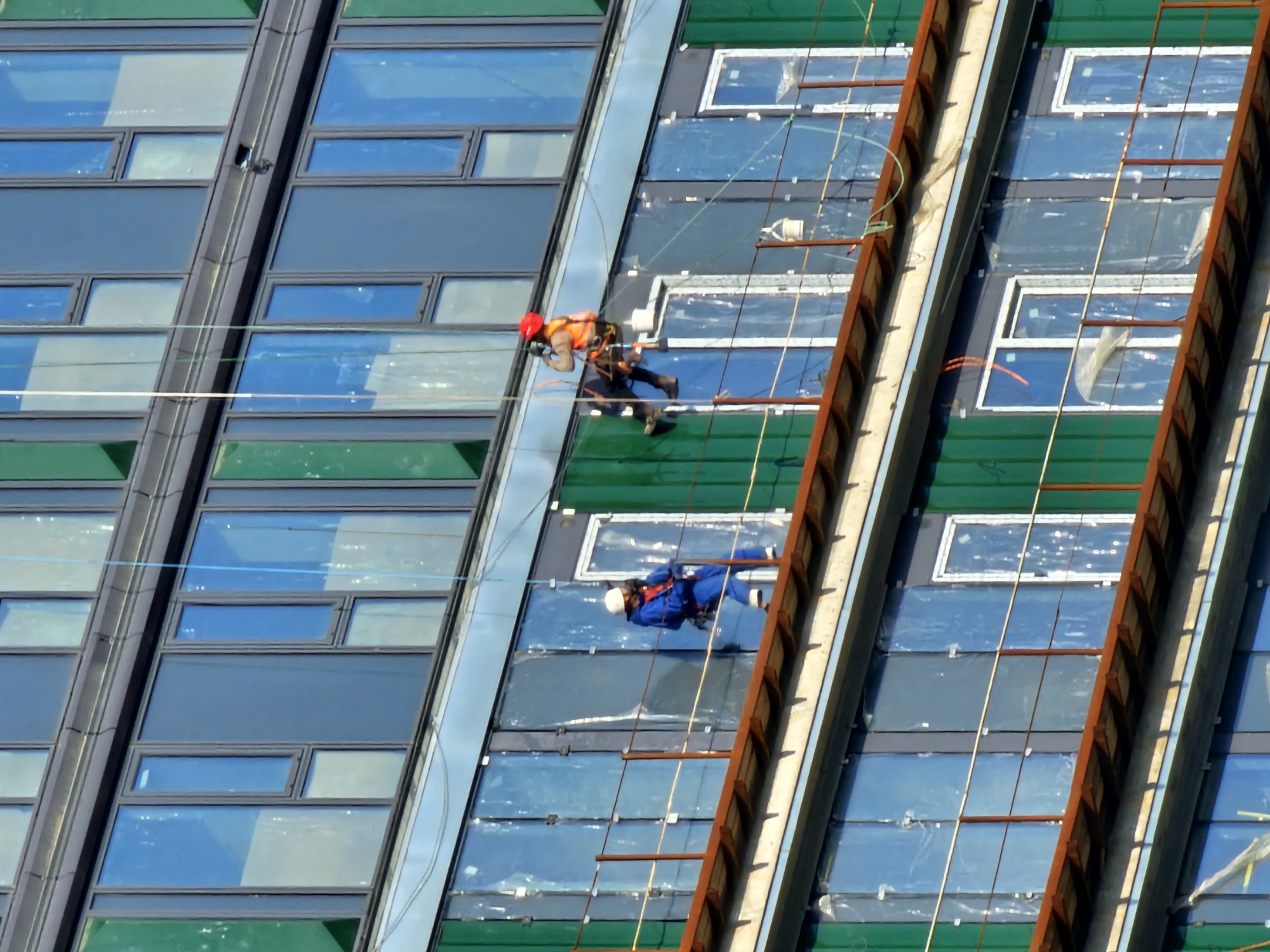 The image shows two workers rappelling down the side of a building, likely in Khimki, Russia, during construction or maintenance. The building has a facade of green and blue-tinted glass windows in a repeating rectangular pattern. The workers are suspended by ropes, one wearing an orange vest and red hardhat, and the other in a blue jumpsuit and white hardhat. A ladder-like metal structure is present on the right side of the image.