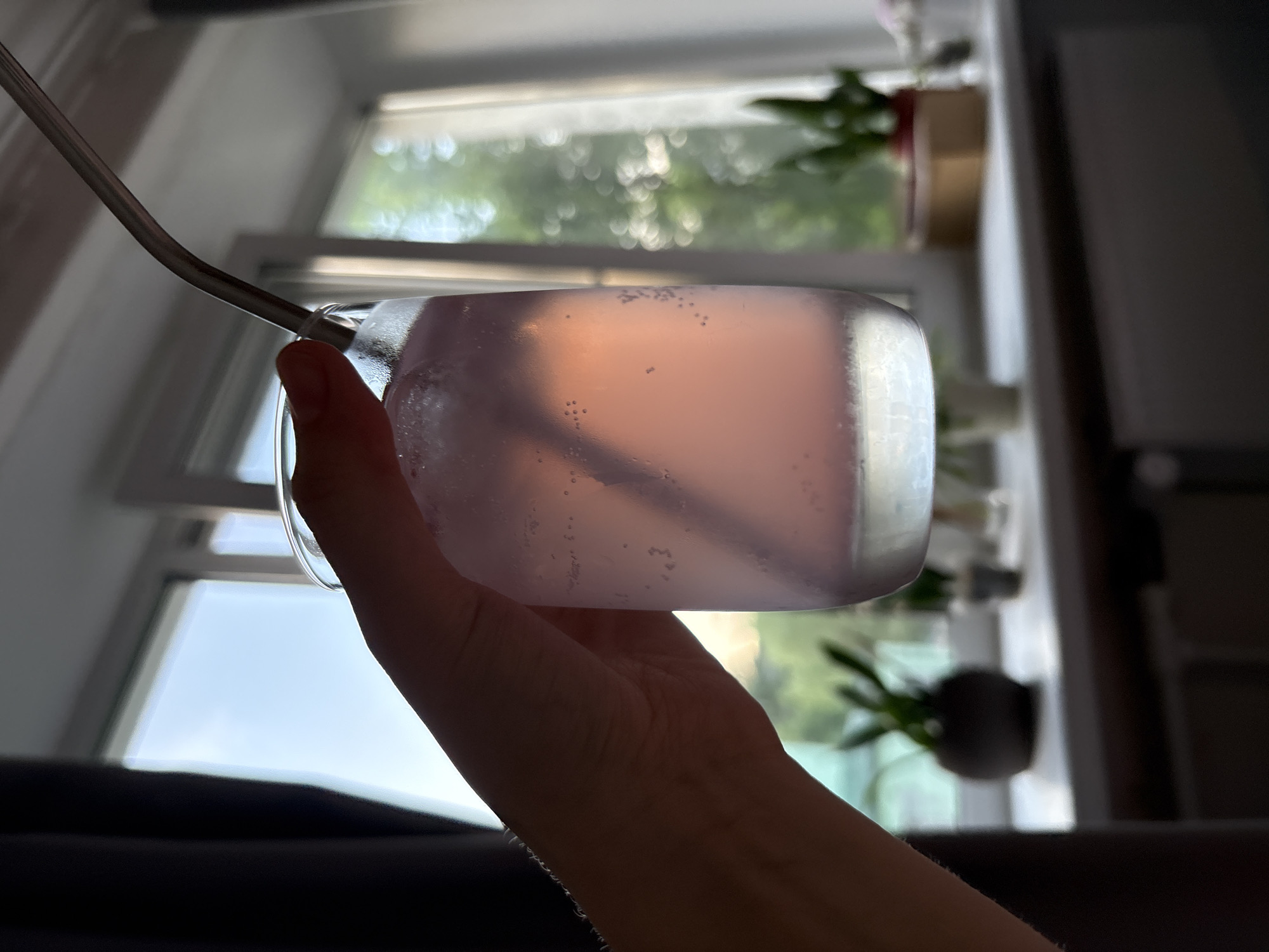 The image presents a close-up, horizontally oriented shot of a hand holding a clear glass containing a light pinkish-purple liquid, viewed from indoors in Saint Petersburg, Russia. The glass, possibly a stemless tumbler, exhibits small bubbles clinging to its inner surface, suggesting a cold or carbonated beverage. A bent metal straw is inserted into the drink.
The person's hand, partially silhouetted, supports the glass from underneath. The background is dominated by a large window through which bright natural light streams in, illuminating the glass and creating subtle reflections. Outside the window, a blurred view of green foliage and trees indicates an outdoor natural setting, characteristic of daytime. The out-of-focus background displays pleasant bokeh circles, consistent with bright, possibly sunny, conditions outside.
Interior elements visible include parts of the window frame and dark, indistinct shapes on the right and bottom, likely furniture or other indoor structures. A small, blurred potted plant can be discerned on a windowsill in the background. The scene conveys a relaxed moment, with someone enjoying a refreshing drink indoors during daylight hours. No text is visible in the image.
