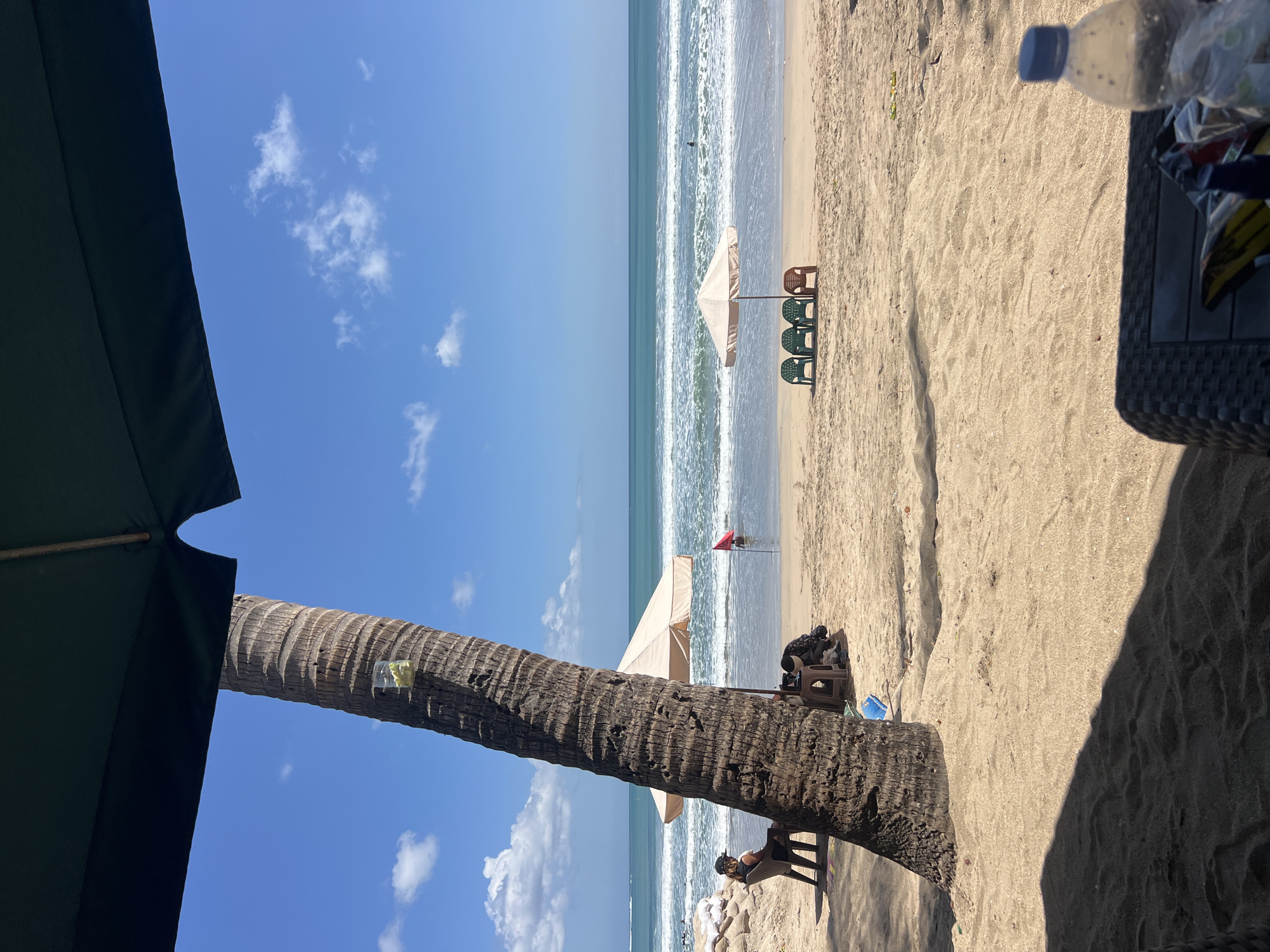 This is a photograph of Kuta Beach in Kuta, Indonesia, on a clear, sunny day. The foreground is dominated by the sandy beach and the shadow of a large umbrella. A thick palm tree trunk is visible on the left side of the frame, with a small clear plastic bag containing yellow items attached to it. In the middle ground, several beach chairs are set up under a beige umbrella near the water's edge. A person is visible in the shallow water further out, near a red flag. The ocean stretches to the horizon under a bright blue sky dotted with white clouds. The overall scene depicts a typical day at a popular tourist beach.