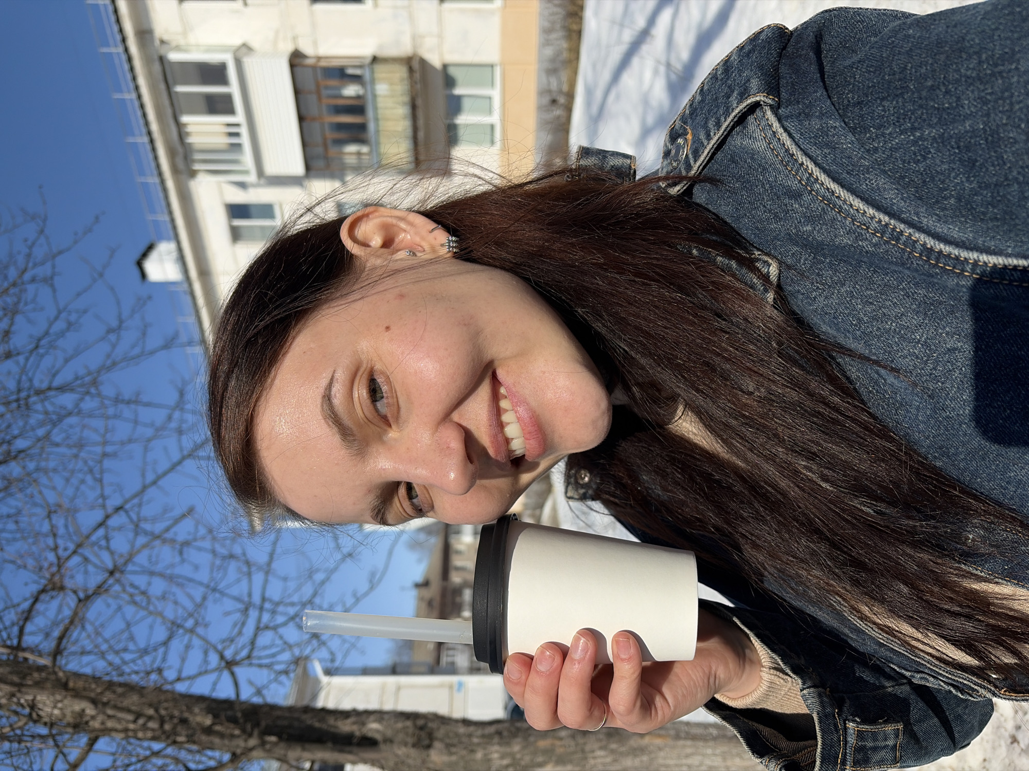 A female subject, appearing to be a young adult, is depicted outdoors in the city of Chaikovsky, Russia. She stands angled slightly to her left, presenting a frontal view with a broad smile and direct eye contact. She has long, dark brown hair, parted centrally and extending over her shoulders. She wears a light-colored turtleneck beneath a dark blue denim jacket. Her left hand holds a white paper cup with a black lid and a clear plastic straw, positioned near her upper chest. Visible on her right ear are multiple silver-colored earrings: a stud in the upper helix, a small hoop in the mid-helix, and a cluster of three small stones in the lobe. A plain gold-colored ring is on her left ring finger. The background features include a bare deciduous tree on the left, snow covering the ground, and a multi-story, light-colored residential building with windows and an enclosed balcony under a clear blue sky. The scene is brightly lit by natural sunlight, casting discernible shadows.