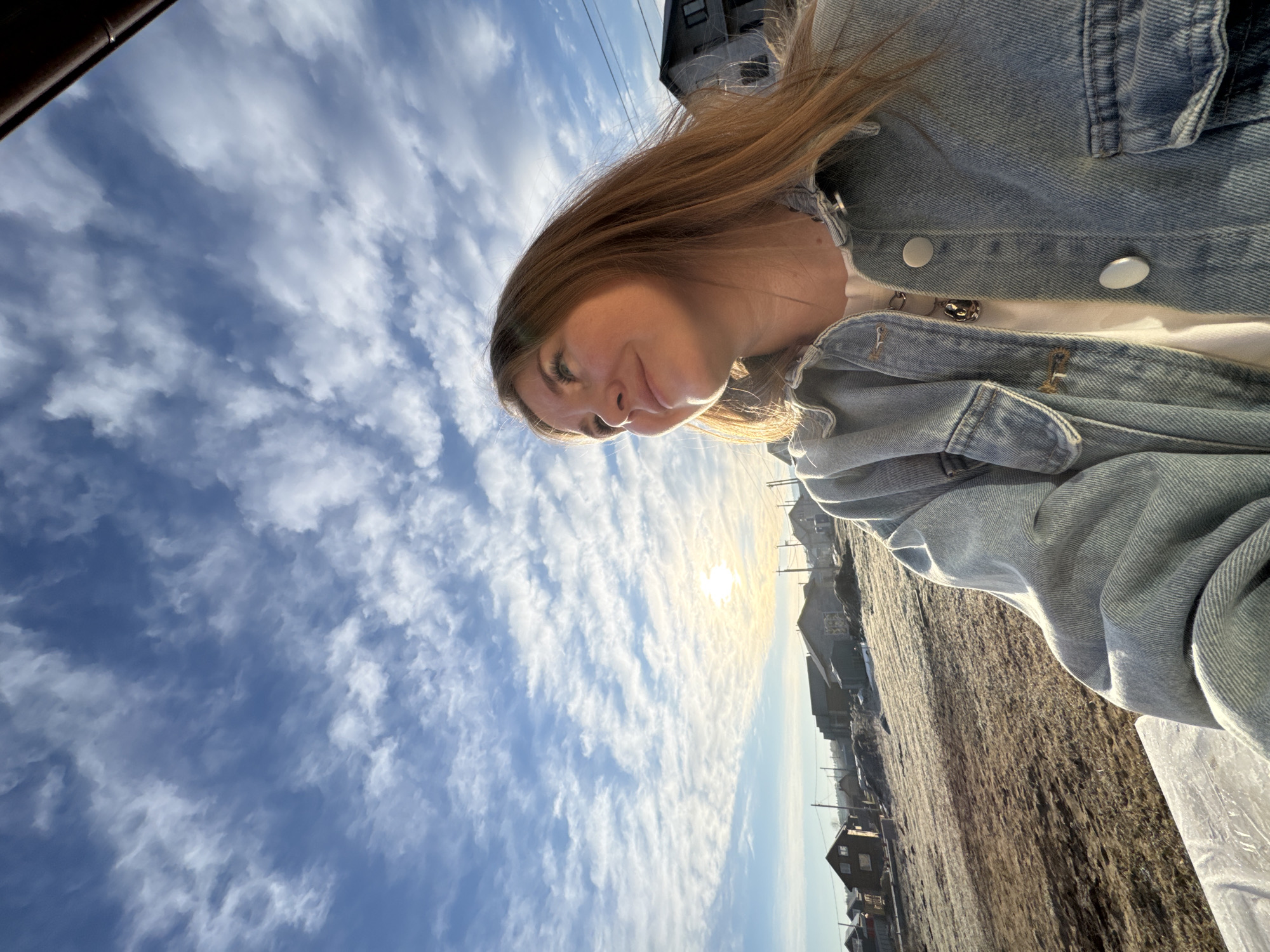 The image shows a young woman in a denim jacket, facing downwards towards the camera. Her hair is light brown. Behind her is a partly cloudy sky, the sun, and a view of a village. The location is identified as being in or near Gornyi (Dvurechenskoe S/P), Russia, given the presence of small, likely residential buildings. The ground appears to be covered in dry grass or dirt. The angle of the shot suggests the picture was taken from a low vantage point.