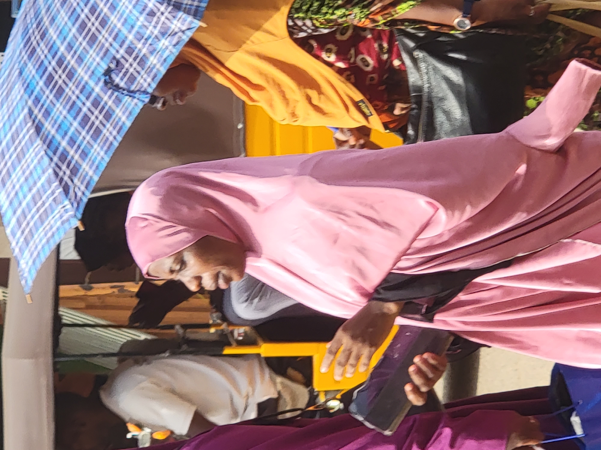 The image captures a bustling outdoor scene, likely a market or street, in Maiburiji, Nigeria, under bright daylight. The composition is dynamic, showing several individuals, mostly from the waist up, and various objects.
The most prominent subject is a woman in the center-right, wearing a flowing light pink hijab and abaya. She faces left, with a neutral to slightly smiling expression, and holds a dark-colored mobile phone in her right hand. To her upper left, another person is partially visible, wearing a vibrant orange long-sleeved shirt with a small, illegible brand tag on the sleeve, and a patterned red and white garment, possibly a skirt or dress. This individual also wears a white-faced wristwatch on their left wrist.
In the upper-left quadrant, a person holds a large blue and white checkered umbrella, open and angled, suggesting protection from the sun or elements. Below the woman in pink, a person in a white t-shirt is visible in the lower-left corner. Various hands are seen throughout the frame, some holding other mobile phones. The background features a yellow structure, possibly part of a stall or vehicle, and wooden planks, indicating a casual, perhaps temporary, setup. The scene conveys a sense of everyday activity in a lively public space.