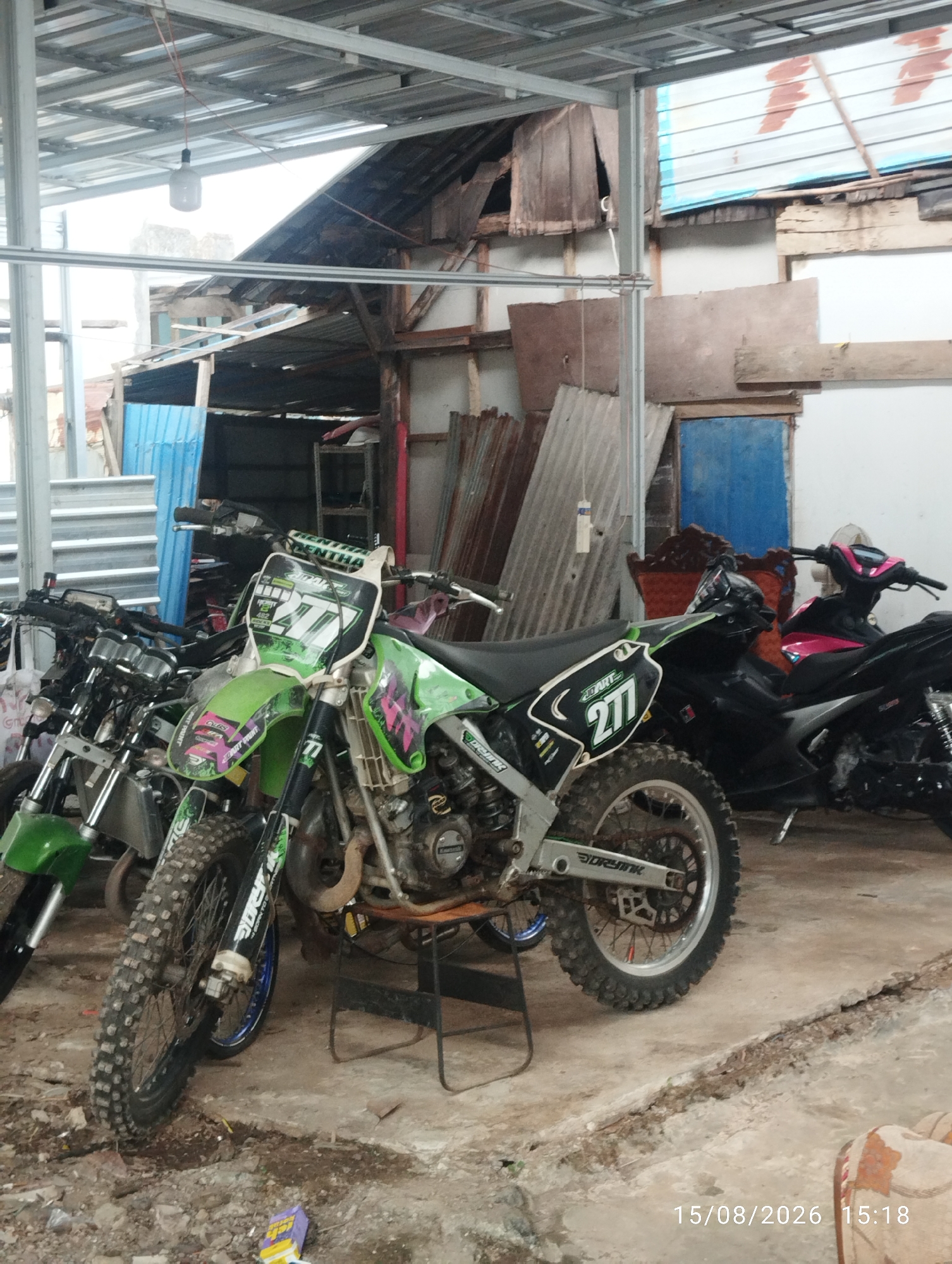 The image shows a collection of motorcycles parked under a metal roof structure. The motorcycles are of various types, including a green dirt bike with the number "277" on its front plate, a black scooter with pink accents, and several other motorbikes with silver and green parts. The environment appears to be an outdoor or semi-outdoor storage area, with corrugated metal walls and a rough concrete floor. Debris and some building materials are scattered around. The background reveals a dilapidated wooden structure and a blue door. The overall scene suggests a functional but unkempt area.