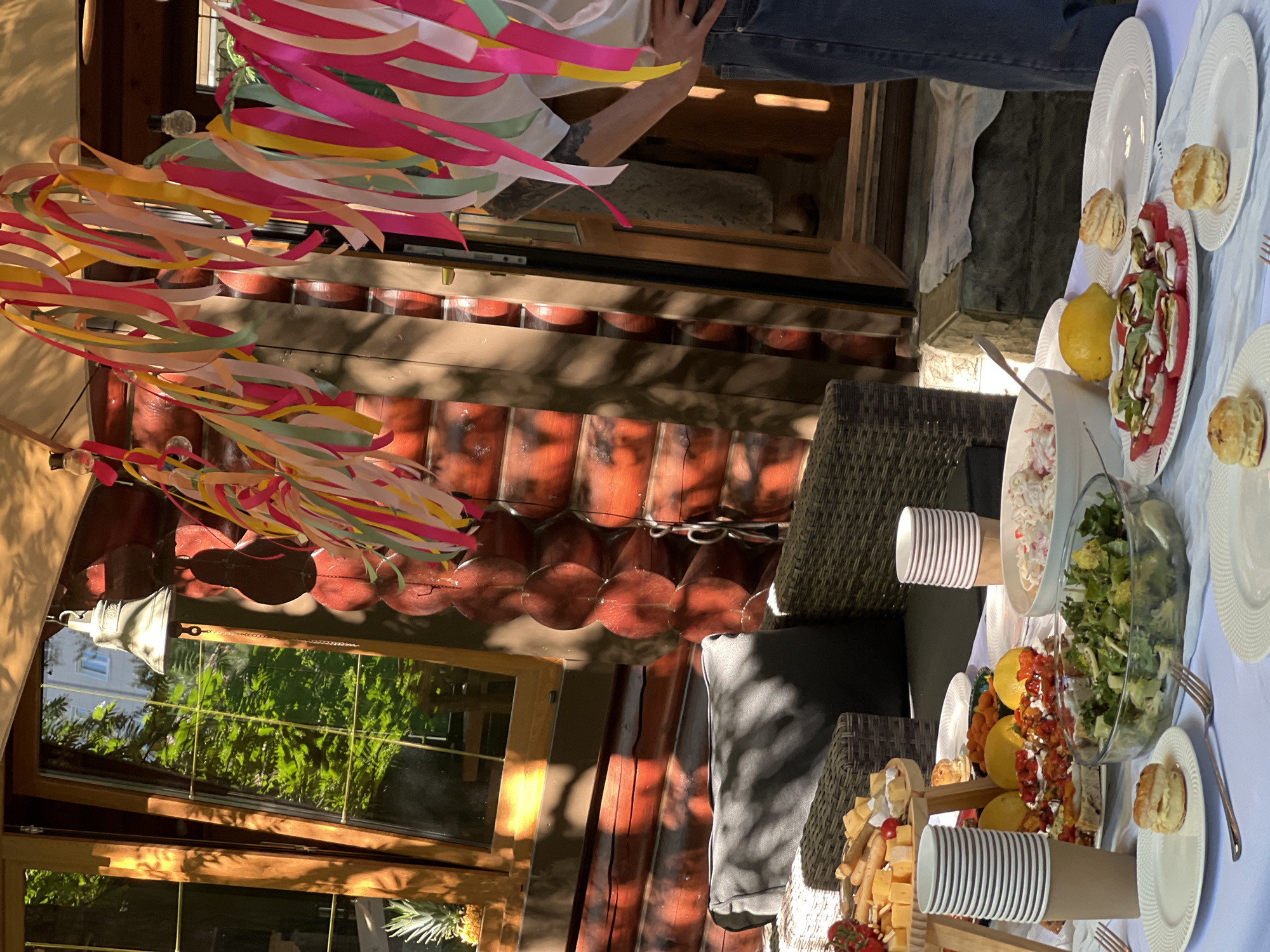 A sun-drenched outdoor gathering is set up on a white tablecloth laid across a table. The table is laden with food, including a cheese and charcuterie board featuring cubes of cheese, breadsticks, and cherry tomatoes. A large glass bowl contains a green salad, next to it a bowl of what appears to be a potato salad. A plate of sliced tomatoes and mozzarella with balsamic glaze is also visible. Two stacks of paper cups and a lemon are also on the table.
In the background, a rustic log cabin structure with a wooden door and windows is visible. Colorful streamers hang from above, adding a festive atmosphere. The environment suggests a backyard or patio setting, possibly in the city of Tomilino, Russia, given the architecture and natural light. A person, partially visible on the right side of the frame, is wearing jeans and a white top, with their arm resting on their hip. The overall scene depicts a relaxed, outdoor meal or party.