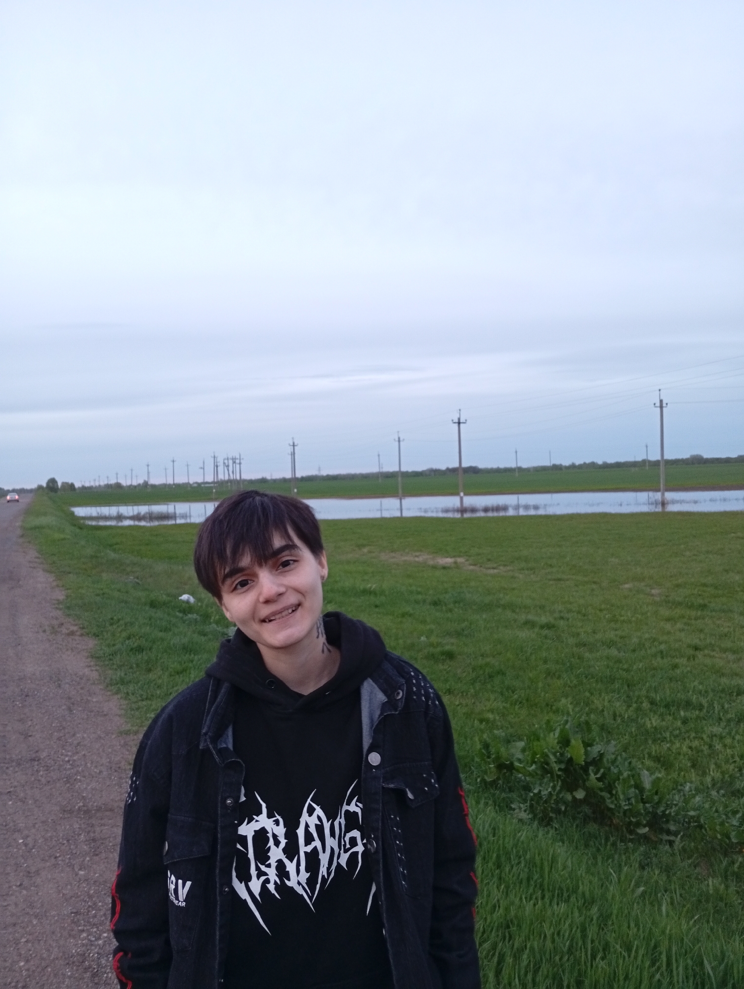 A young person stands on a dirt road in Azyakul', Russia, with a flooded field behind them. The person is smiling and wearing a black hooded sweatshirt with a white graphic design and a black denim jacket with red stripes on the sleeves. Power lines run along the field. A car is visible in the distance on the road. The sky is overcast.