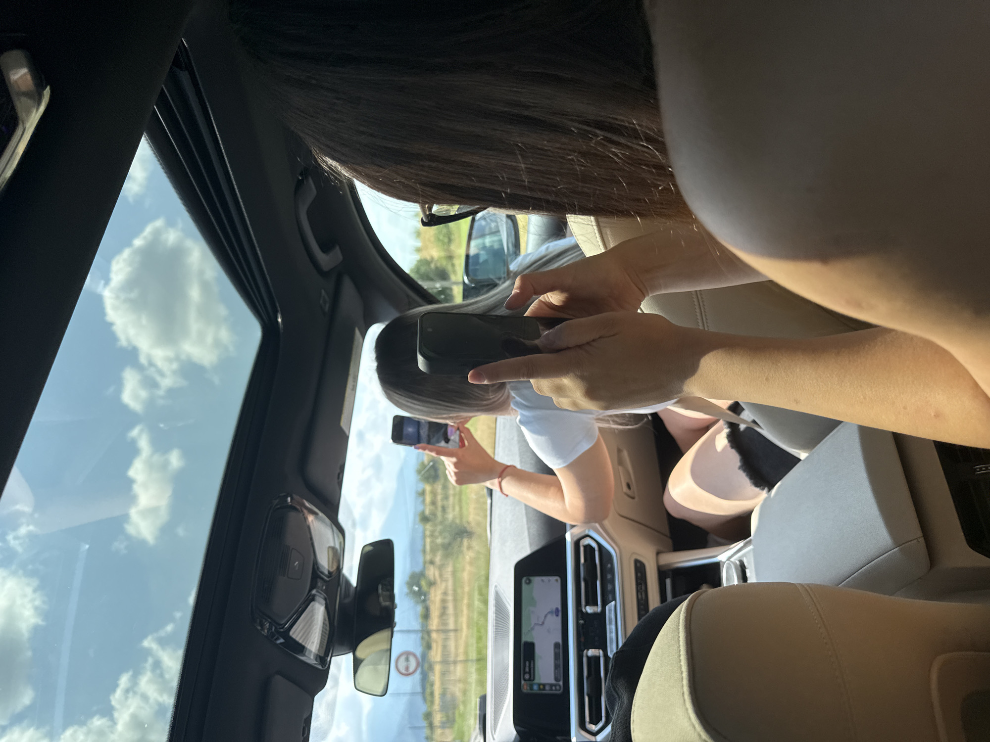 In the city of Cârțișoara, Romania, two women are inside a car, with a sunroof open to a bright, cloudy sky. The woman in the passenger seat is taking a photo with her smartphone of the scenery outside. The driver, facing away from the camera, is also holding a smartphone, possibly taking a photo or recording a video. The car's interior includes a navigation screen displaying a map, a rearview mirror, and the beige leather seats. The exterior view shows a road with fields and mountains in the distance.