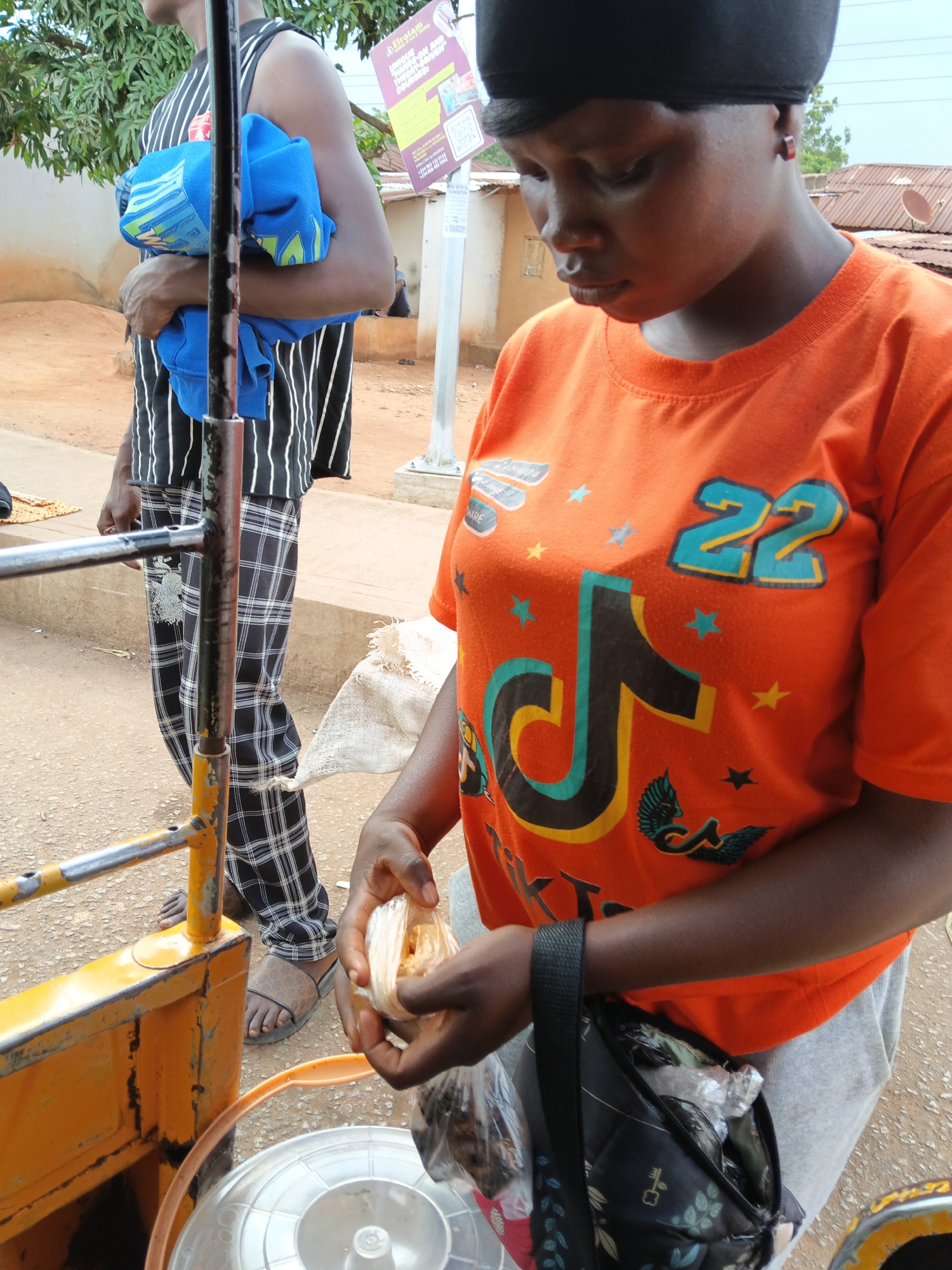A person in an orange t-shirt with a TikTok logo and the number "22" is holding a clear plastic bag with some food items inside. They are wearing gray sweatpants and have a black bag slung over their shoulder. To their left, another person is standing wearing a black and white striped sleeveless shirt and plaid pants, holding a blue garment. They are both near a yellow cart with metal railings. The ground appears to be dirt or gravel, and there are some buildings and trees in the background. A sign with text is visible behind the person in the orange shirt. The lighting suggests it is daytime, possibly outdoors in a warm climate.