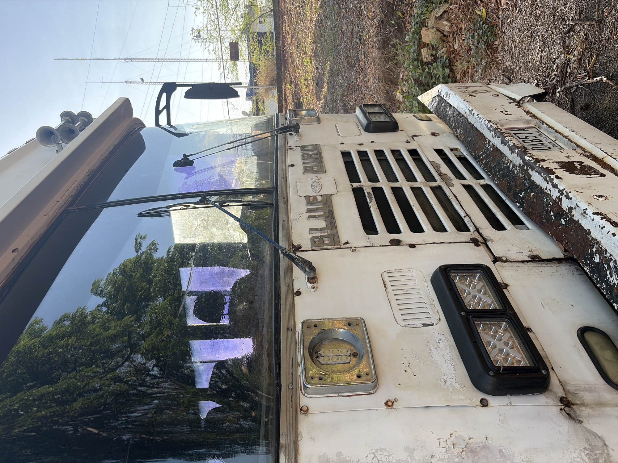 An overhead, angled view captures the front section of a weathered white vehicle, likely a truck or bus, situated outdoors in Ciudad Guayana, Venezuela. The vehicle's paint is peeling and rusted in various spots, indicating age or disuse.
The prominent front of the vehicle features a large windshield reflecting lush green trees and a clear blue sky. Two windshield wipers are visible, one resting on the glass and the other slightly lifted. On the roof above the windshield, a pair of chrome air horns are mounted.
The front grille, featuring vertical slats, is visible on the right side of the image. Above the grille, metal lettering spells out "BIRD" and "BULL," possibly parts of a manufacturer's name, with an emblem or logo positioned between them. To the right of the grille, two light fixtures are present: one is an empty, circular housing with small internal lights, and the other is a rectangular black unit containing two rows of modern LED lights.
Further to the right, a white and rusted bumper or body panel extends outwards, displaying a license plate. The visible characters on the license plate are "DAB57", with "DISTRITO CAPI" (likely "Distrito Capital") printed below it.
The background shows a bright, clear sky with tall structures resembling power lines or transmission towers. The ground around the vehicle is covered with dry, reddish-brown leaves and foliage. The scene suggests a daytime setting with bright sunlight. The overall impression is of a robust vehicle that has seen considerable wear and tear, possibly parked in an industrial or semi-rural area.