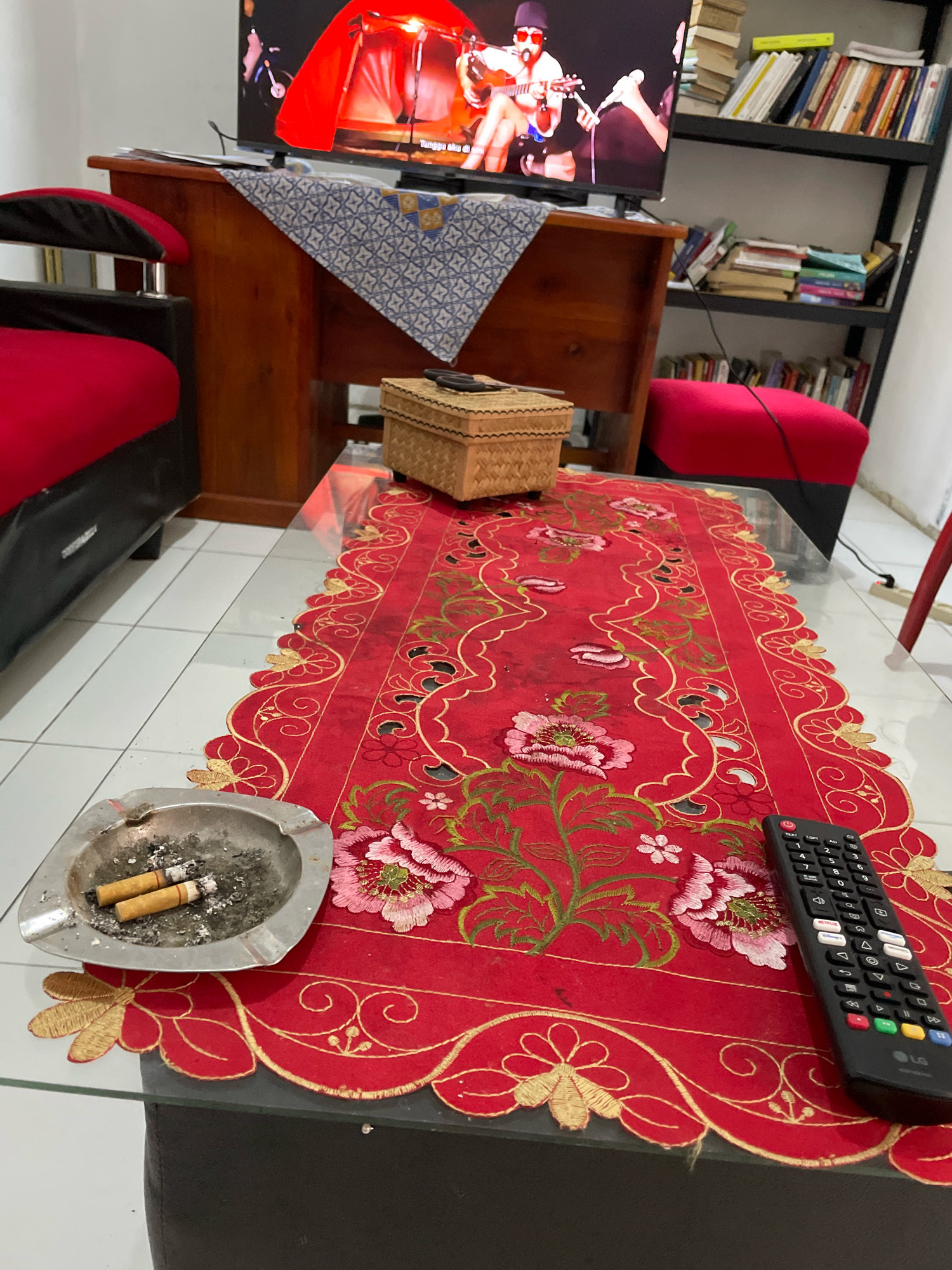 The image depicts a living room scene in Kagungan Ratu, Indonesia. A glass-top coffee table is covered with a red embroidered table runner featuring floral motifs in pink, green, and gold. On the table, an ashtray containing two cigarette butts sits to the left, next to a woven tissue box. A black remote control rests on the right side of the table runner. In the background, a television screen displays a musical performance. To the left of the television, a wooden cabinet supports the television and a blue and white patterned cloth. A red armchair with black accents is partially visible on the far left. A bookshelf filled with books stands to the right of the television, with a red pouf placed in front of it. The floor is tiled with white and gray squares.
