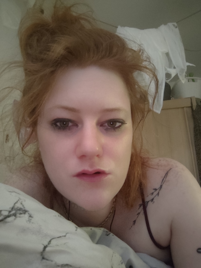 A pale-skinned person with disheveled red hair and dark eyeliner gazes at the camera. Their hair is piled on top of their head. They are lying on a pillow with a gray marble pattern. The individual wears a thin, gold chain necklace and a burgundy top. A small tattoo of a leafy branch is visible on their left shoulder. In the background, plastic bags hang from a line, and a beige cabinet stands to the right.