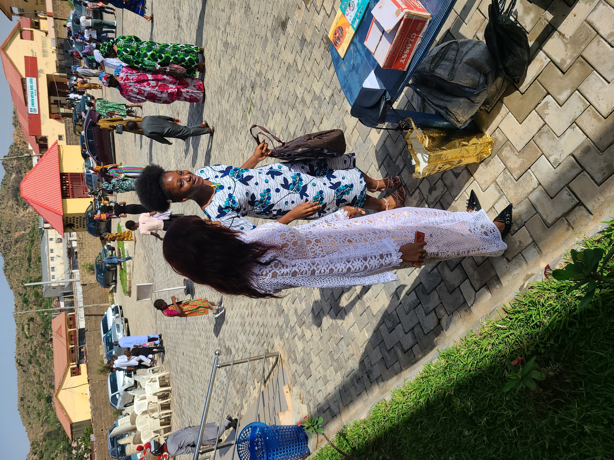 The image depicts an outdoor scene featuring two women standing next to a table with various items on it. The woman on the left is wearing a blue and white floral dress and holding a brown handbag. The woman on the right is wearing a white lace dress and dark shoes.
Behind them is a crowd of people, some wearing colorful traditional clothing. In the background, there are buildings, cars, and a hillside.
The ground is paved with patterned stones. There is green grass visible on the bottom right corner of the image.
The location is in the city of Unknown, Unknown.
