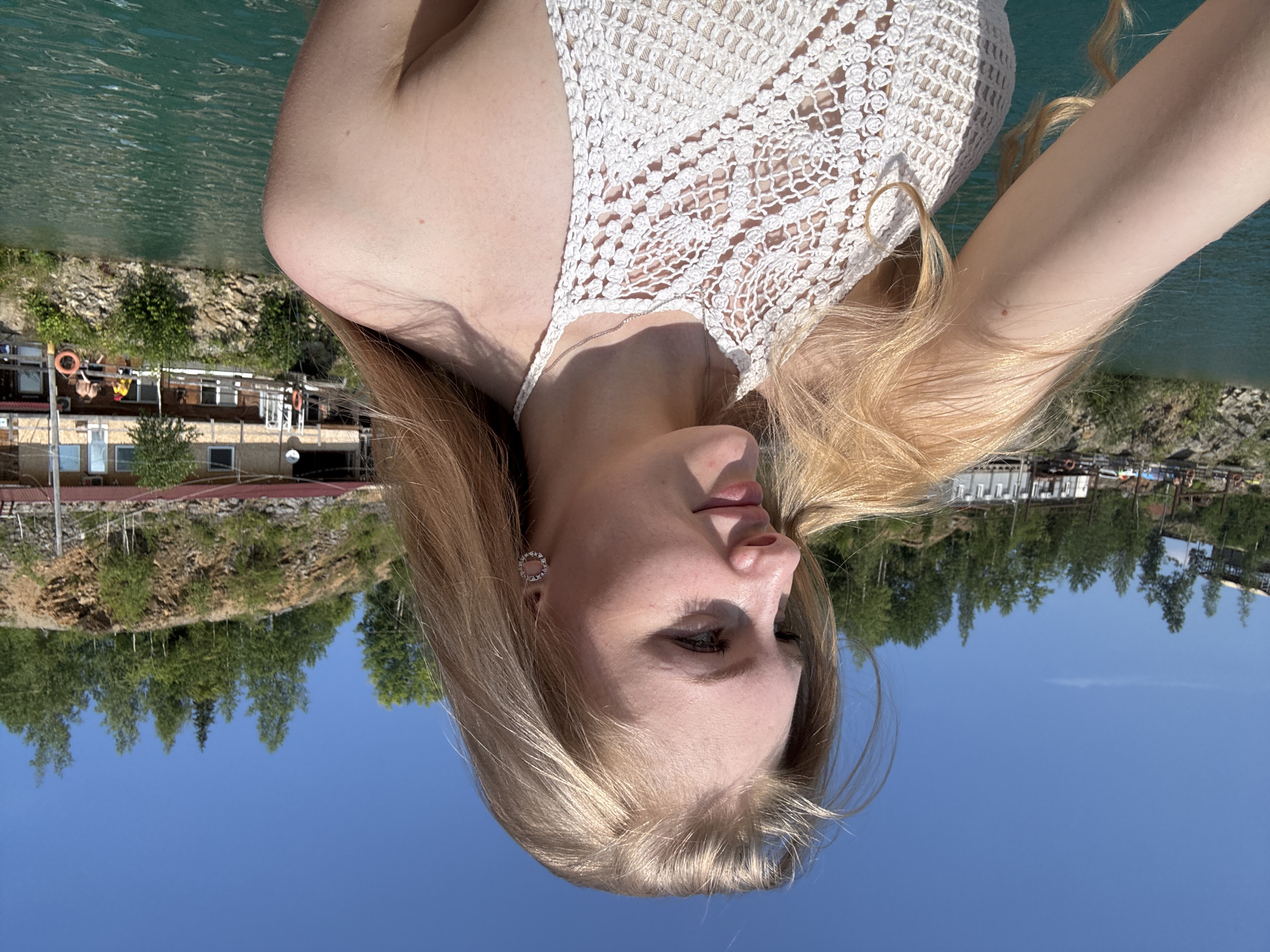 A young woman with blonde hair is in the foreground, facing to the left, with a blue sky and a rocky hillside in the background. She is wearing a white crocheted halter top and a silver necklace. The water is a bright turquoise color, and there are buildings and trees on the hillside. The scene appears to be a quarry lake in Karyer Izvestnyak, Russia. Several people are visible on a dock in the background, and some are in the water. The overall impression is a sunny day at an outdoor recreational area.