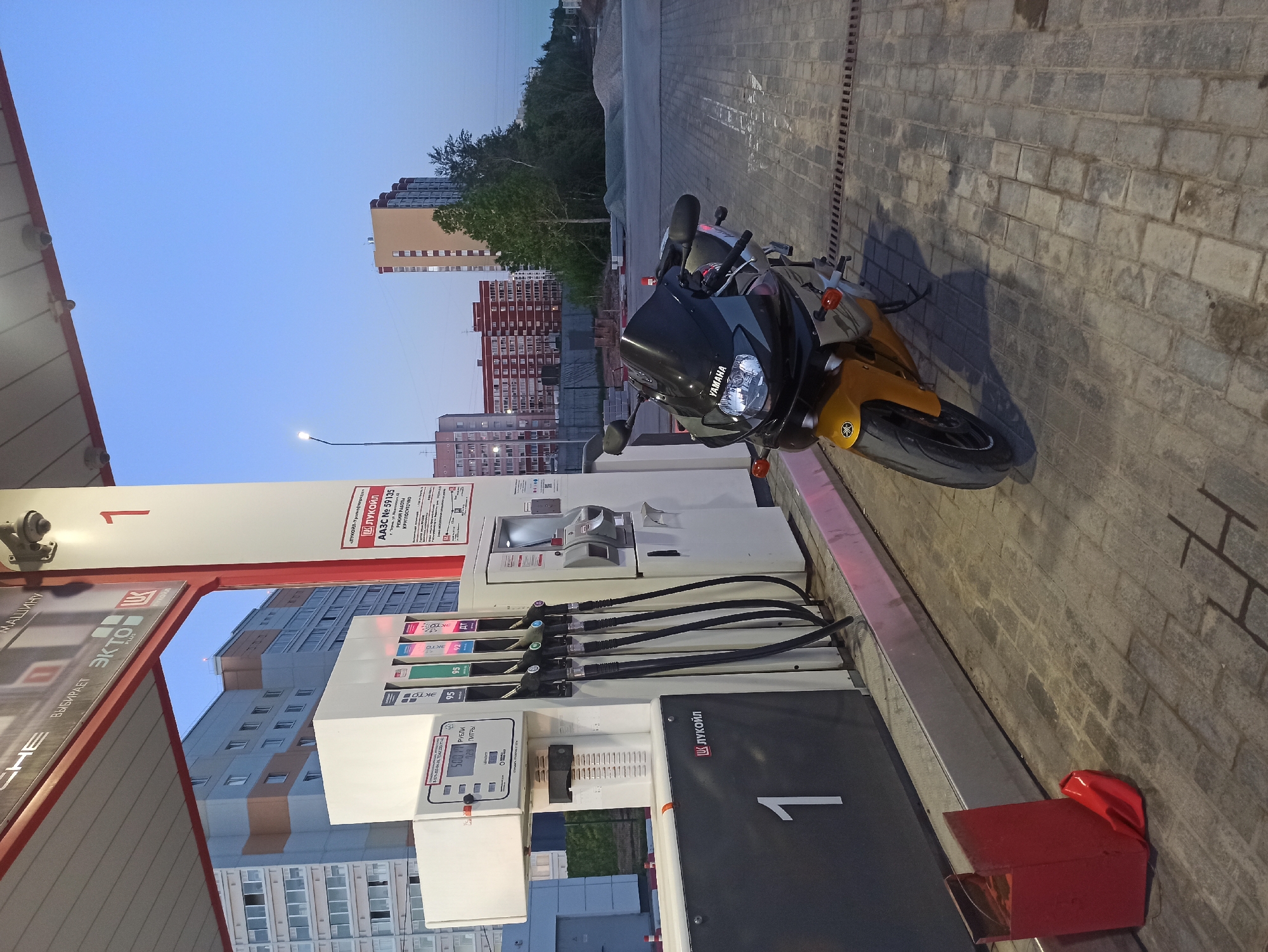 A motorcycle is parked at a gas pump in an urban area. The motorcycle is a Yamaha and appears to be yellow and black. The gas pump is white with black and red accents, and the signage identifies it as a Lukoil station, specifically AАЗС No. 59135, which is open 24 hours. The pump is displaying a sum of 500 rubles. In the background, there are several apartment buildings of varying heights and colors, indicating an urban setting. The sky is a pale blue, suggesting it's either early morning or late evening. The ground is paved with grey rectangular stones.