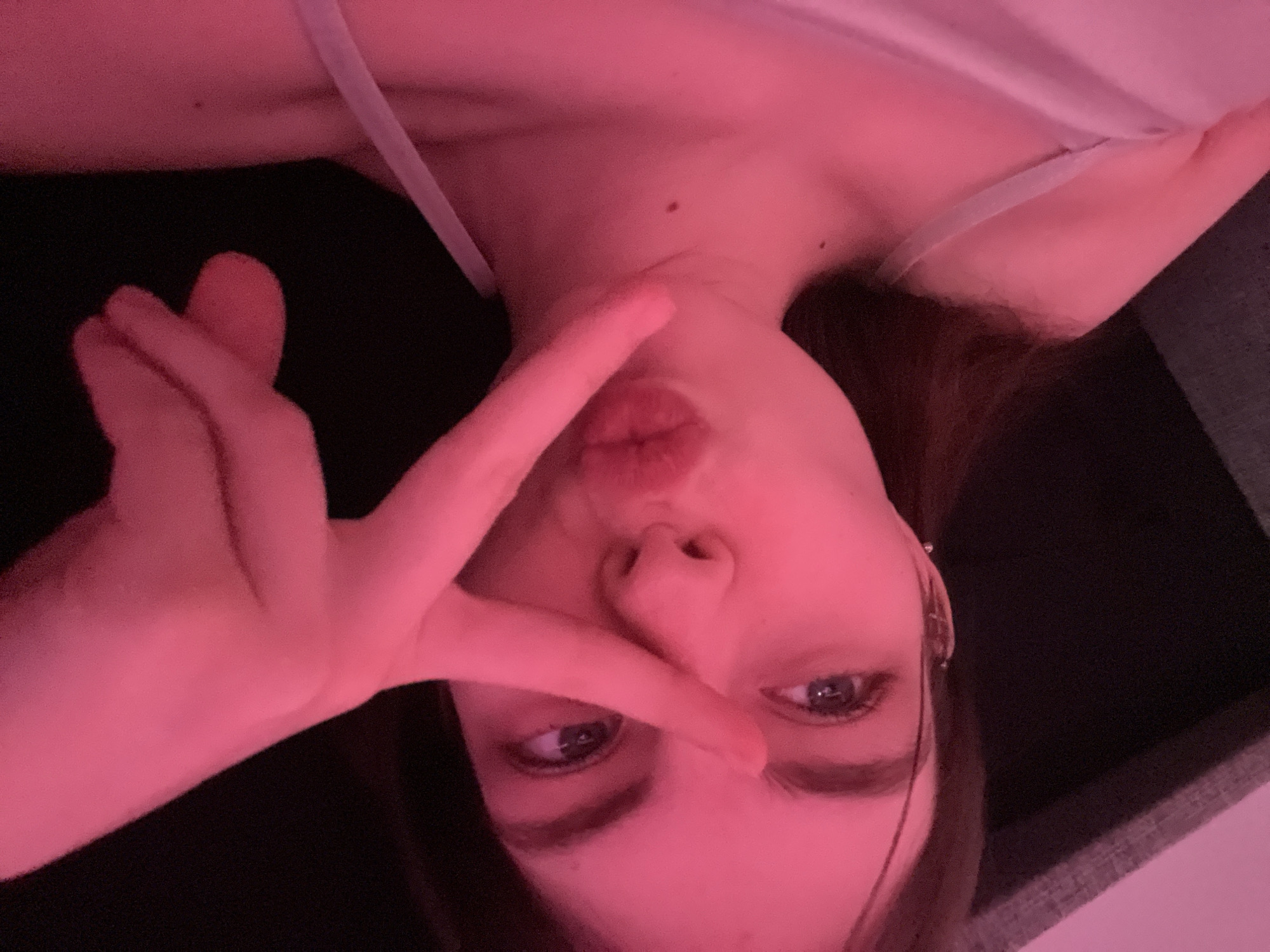 A young woman is posing for a selfie. Her face is oriented in a way that suggests she is holding the camera above her head. She has light skin, dark hair, and blue eyes. She is making a kissy face, and her left hand is raised, forming a "V" sign over her face. She appears to be wearing a light-colored top with thin straps. The background is dark, but it appears she is in an indoor setting. There are no indications about Saint Petersburg, Russia.