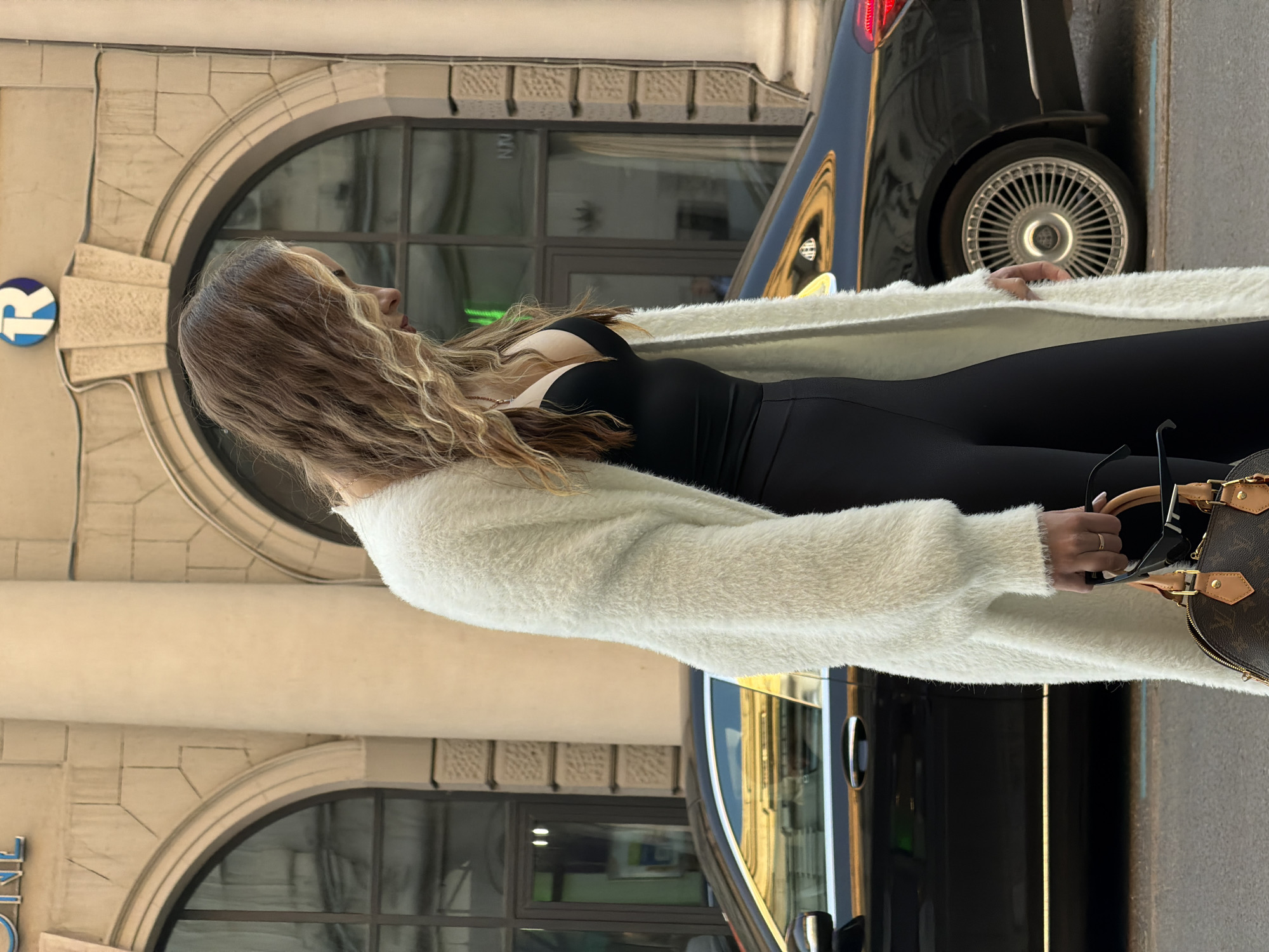A woman with brown hair and a white sweater is standing outside on a city street. She is wearing a black jumpsuit and holding a brown Louis Vuitton bag and black sunglasses. A black car is parked beside her. The building behind her has an arched window and a sign with the letter "R" on it. The sign indicates she is likely in Moscow, Russia.