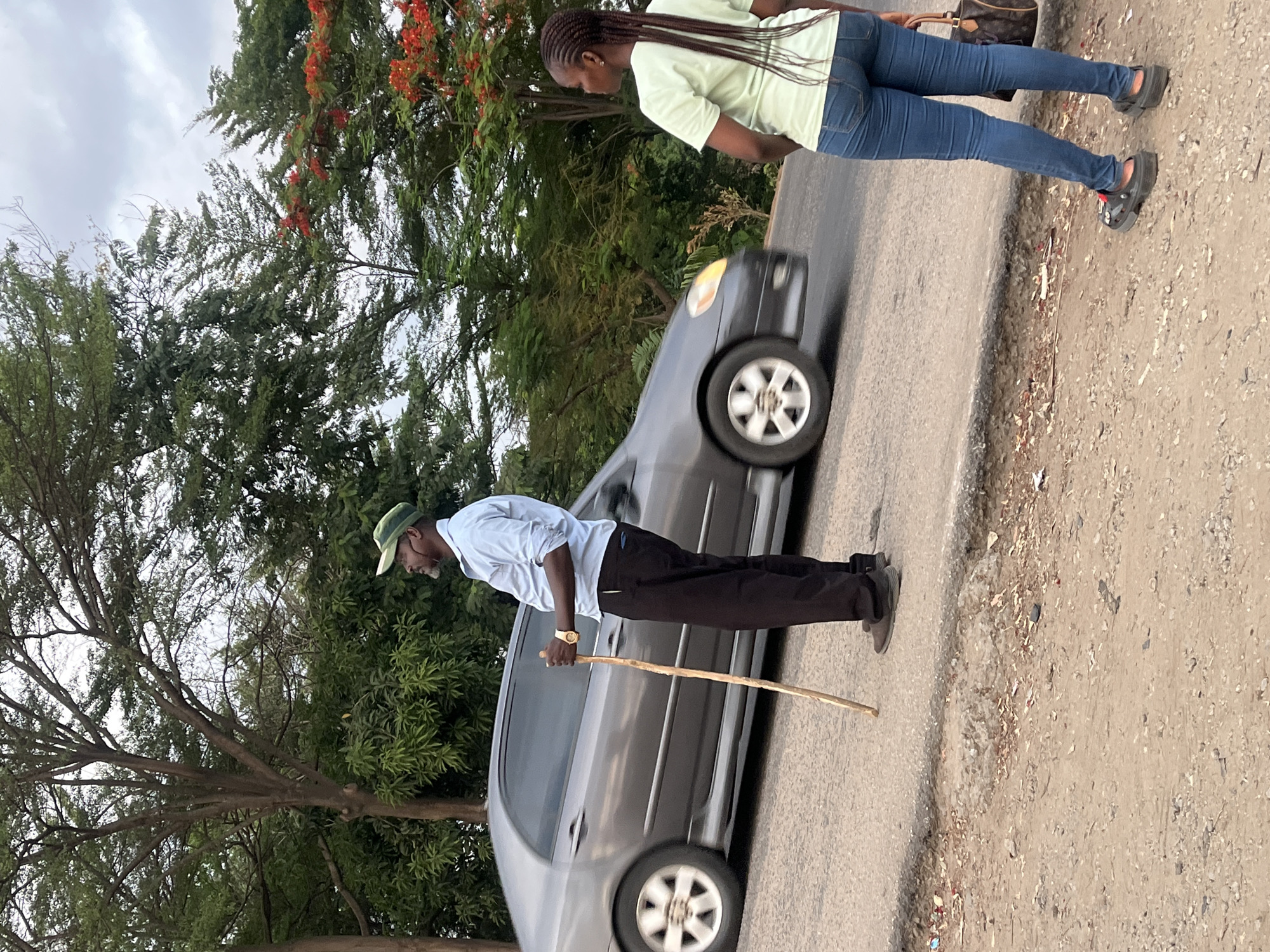 A medium shot, from ground level, of a grey four-door sedan car driving on a narrow road in Karmajiji, Nigeria. An elderly Black man in a light blue button-down shirt, black pants, a green cap, and holding a walking stick stands next to the driver's side of the vehicle on the left side of the road. A young Black woman in a light green t-shirt, blue jeans, and black sandals walks ahead of the car on the right side of the road, carrying a brown handbag. Trees and plants are visible on both sides of the road. The sky is overcast.