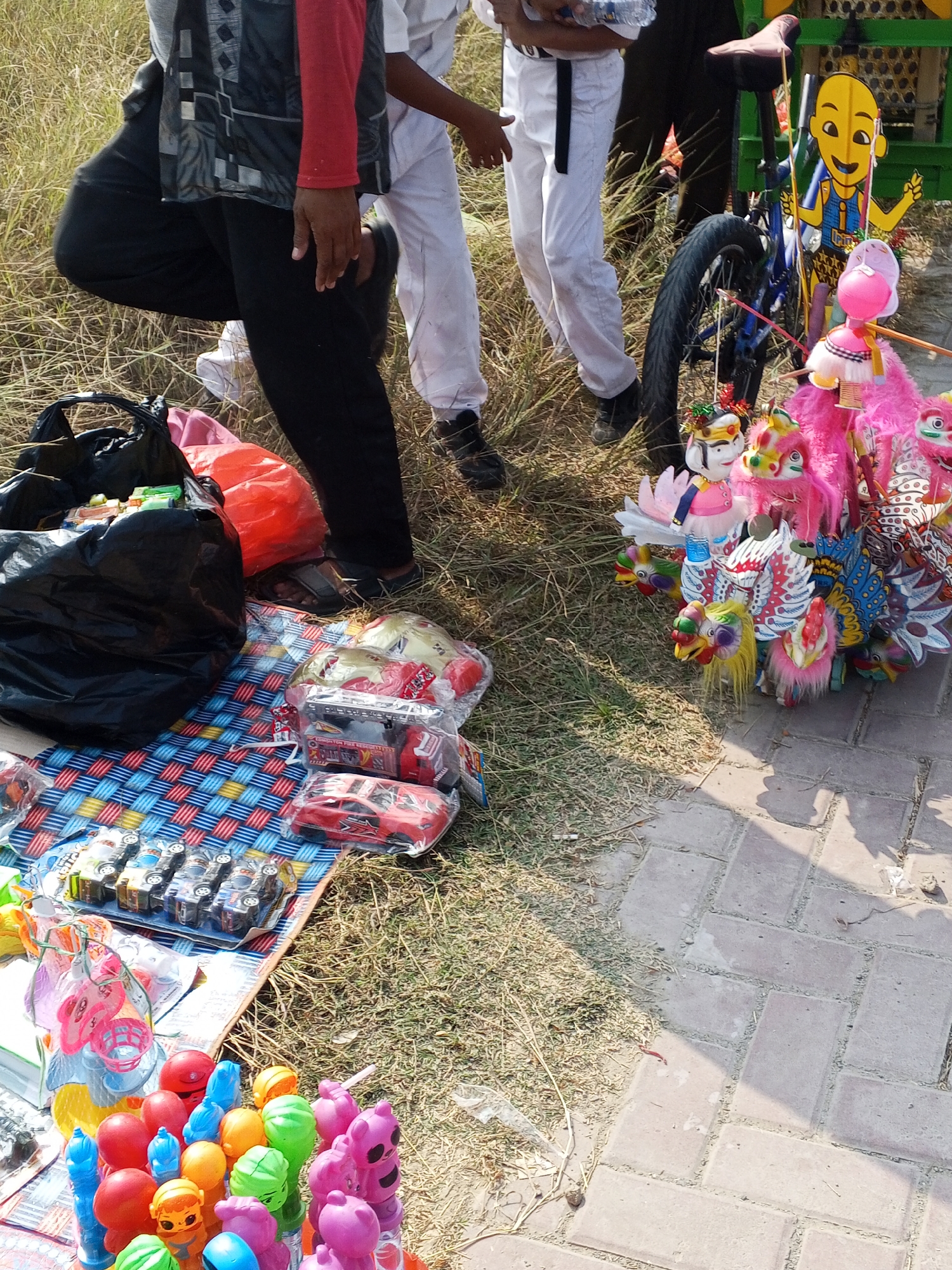 The image captures a street vendor scene, likely in the city of Ranjeng, Indonesia. The foreground is dominated by a display of colorful toys, including what appear to be toy cars, dolls, and novelty items like spinning windmills and character figures with vibrant headdresses. The toys are arranged on a patterned mat and on the ground. Behind the toys, a person in dark pants and a red and black patterned shirt is partially visible, tending to the merchandise. To the right, a bicycle with a bright yellow, cartoonish figure attached is propped up. In the background, two individuals in white clothing are present, one holding a plastic bottle. The environment consists of dry grass and a paved walkway made of brick pavers. The overall impression is that of a lively outdoor market or roadside stall.