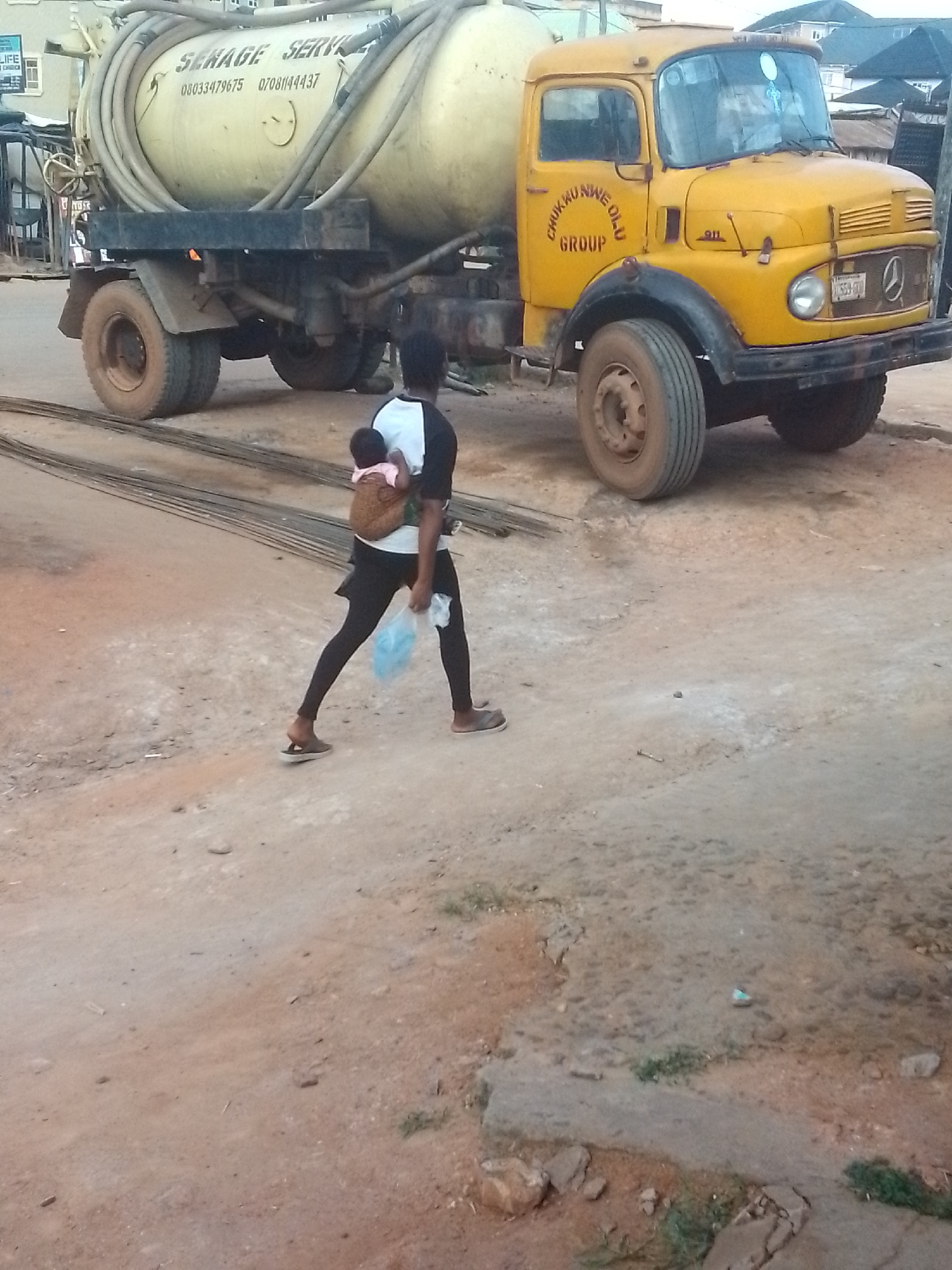 The image depicts a woman carrying a baby on her back, walking on a dirt road in Nkpor, Nigeria. She is wearing a black and white shirt, black leggings, and sandals, and is holding a blue plastic bag in one hand. Next to her is a large, yellow sewage truck with the words "SEWAGE SERVICE" and phone numbers written on the tank. The truck also has "CHUKWU NWE OLU GROUP" and "911" painted on the side. The road is uneven and partially covered with exposed pipes. The general environment suggests a developing urban area.