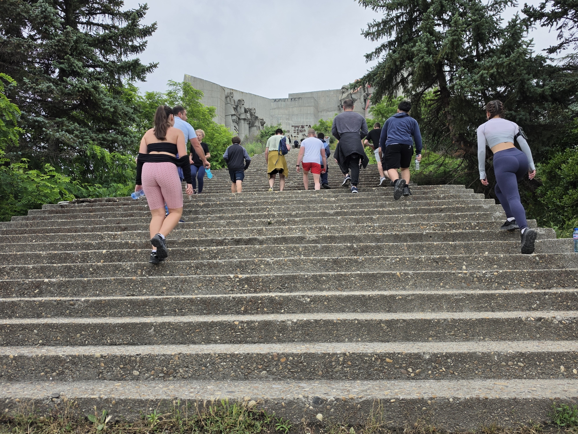 The image depicts a group of people ascending a wide, long, concrete staircase leading towards a monumental structure in Varna, Bulgaria. The monumental structure appears to be a memorial or monument, with large concrete slabs and sculptures visible. Graffiti, including the words "CALVIN KLEIN," is spray-painted on the facade of the monument. The staircase is made of rough, gravel-infused concrete and is flanked by lush green trees on either side.
In the foreground, several individuals are seen climbing the stairs. A woman in pink athletic shorts and a black bandeau top is in the lower left, holding a blue water bottle. To her right, a man in a light blue shirt and dark shorts is also ascending. Further up the stairs, a diverse group of people, including men and women of various ages, are climbing. Some are dressed in athletic wear, suggesting they might be exercising or hiking. A woman in a light purple top and dark blue leggings is prominently featured in the upper right, also climbing the stairs. The sky is overcast, contributing to a diffused light. The environment suggests an outdoor, possibly historical or commemorative site, popular for physical activity.