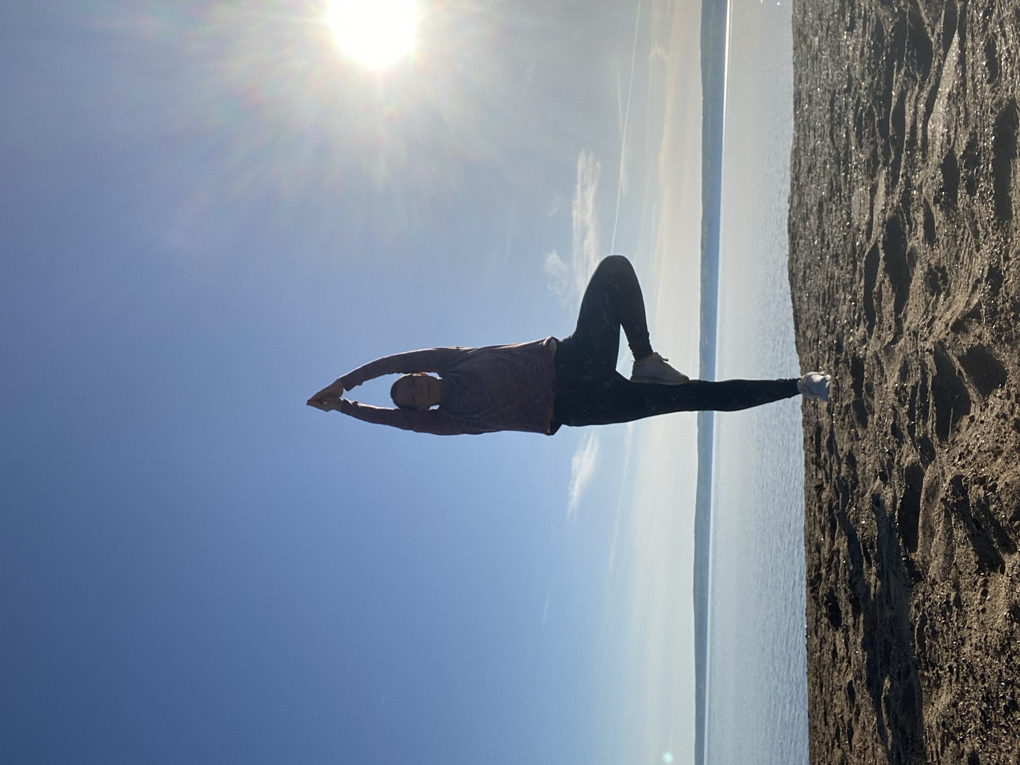 The image shows a woman performing a yoga pose outdoors in Chaikovsky, Russia. She lies on her back on a sandy surface next to a body of water, with her legs extended to the right and slightly elevated, and her arms stretched above her head. She is wearing dark leggings, light-colored sneakers, and a long-sleeved shirt. The sky above is a clear blue with a bright sun near the top of the frame. The body of water appears calm and stretches towards the horizon. The sandy surface occupies the right side of the image and appears coarse.