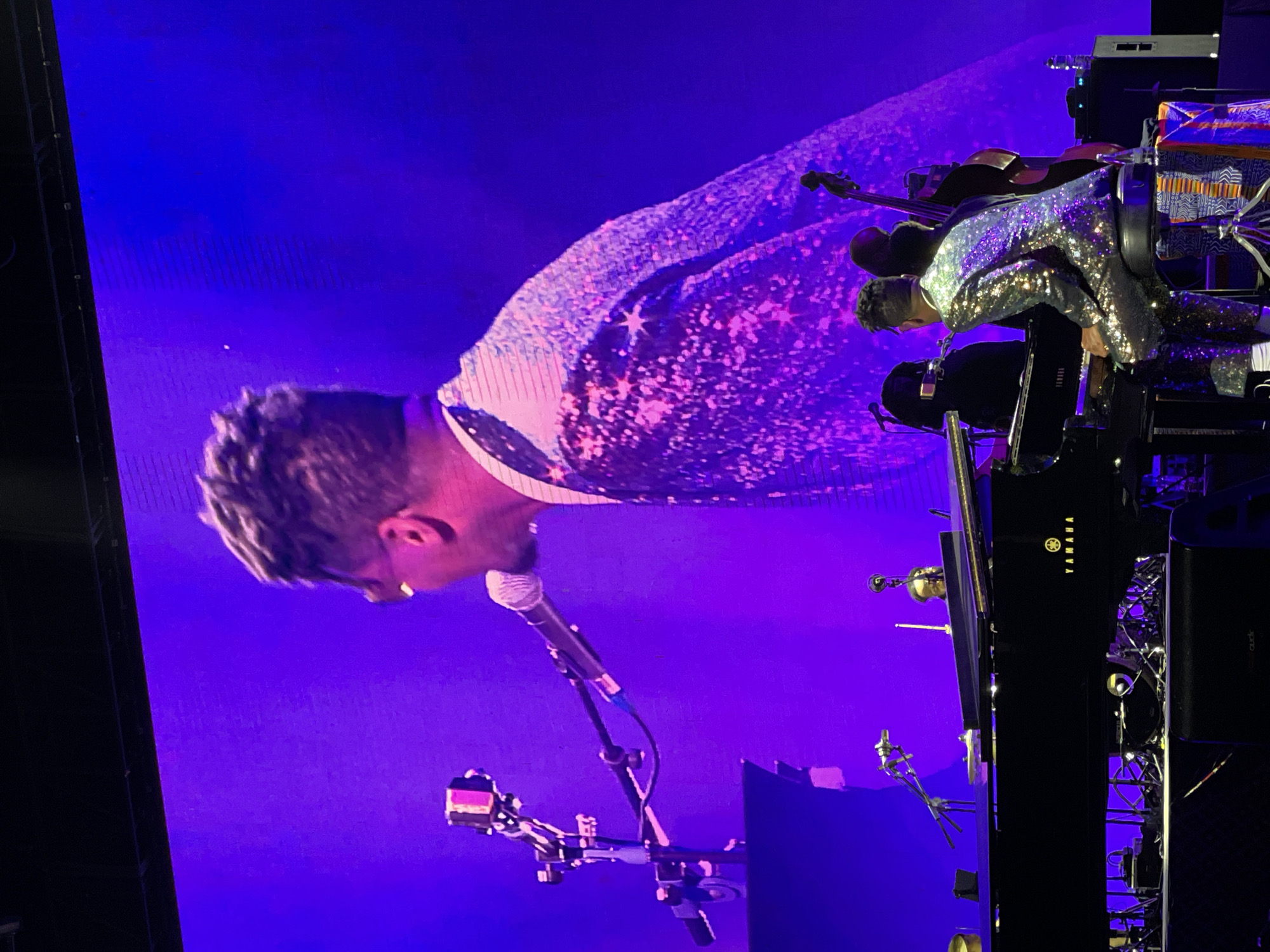 A man is performing on stage, likely at a concert, in Salembaran, Indonesia. The man is wearing a sparkling suit and is positioned at a Yamaha piano. He is leaning toward a microphone, presumably singing or speaking. A screen behind him displays a large, brightly lit image. There are microphones and other musical equipment visible around the piano. The lighting is primarily blue and purple, creating a vibrant concert atmosphere.