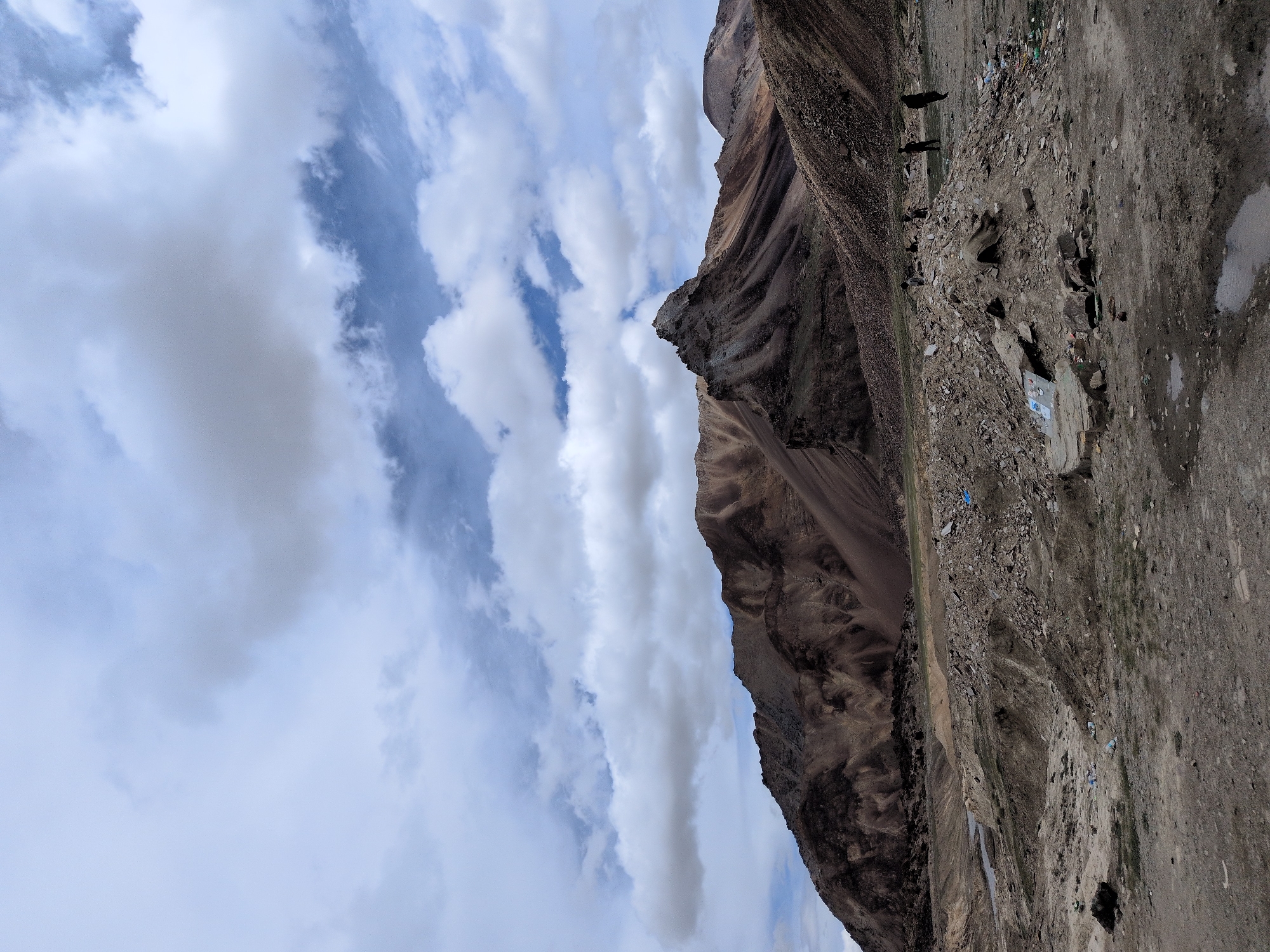 In Killingsarai, India, a vast, arid landscape is dominated by rugged, brown mountains under a partially cloudy sky. The foreground is a desolate, rocky terrain with scattered debris, including plastic bottles. Several figures, appearing as dark silhouettes, are dispersed across the middle ground, some standing near a pile of rocks and others walking. The mountains in the background exhibit a layered, striated appearance, with sharp peaks and eroded slopes. The sky is a mixture of pale blue and white clouds, with some appearing heavy and grey.