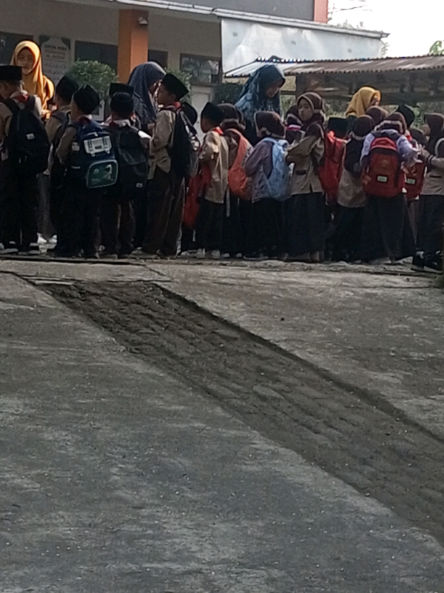 In Krasak, Indonesia, a group of students, predominantly boys wearing traditional Muslim attire, stand in a courtyard. They are dressed in light-colored shirts and dark pants or skirts, many with matching caps. Most of the students are carrying backpacks, varying in color and design, including blue, red, and black. Some students are wearing hijabs, suggesting a mixed-gender group or that some older students are accompanying younger ones. The students appear to be waiting or gathered, possibly for a school activity or assembly. The environment suggests an outdoor setting, likely a schoolyard, with a building in the background. The ground is paved with asphalt, showing signs of wear and tear, with a dark, textured pattern suggesting a shadow or a change in the paving material. The lighting is somewhat subdued, indicating it might be early morning or late afternoon. The overall impression is of a typical school day scene in an Indonesian community.