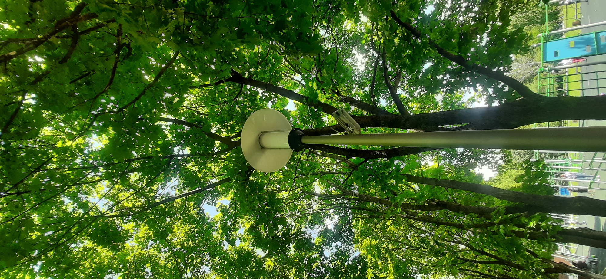 This is a low-angle, vertical shot of a park in Yessentukskaya, Russia, on a sunny day. The foreground is dominated by a tall, metallic lamp post with a white, circular lamp fixture at the top. Attached to the lamp post, slightly below the fixture, is a security camera. The background is filled with the lush, green canopy of mature trees, with sunlight filtering through the leaves, creating dappled light patterns. In the middle ground, and slightly blurred due to the focus on the lamp post and trees, is an outdoor exercise equipment area. Several people are visible in this area, some standing near the equipment, others walking. The ground appears to be a light-colored, soft surface, possibly rubberized, commonly found in children's playgrounds or exercise zones.