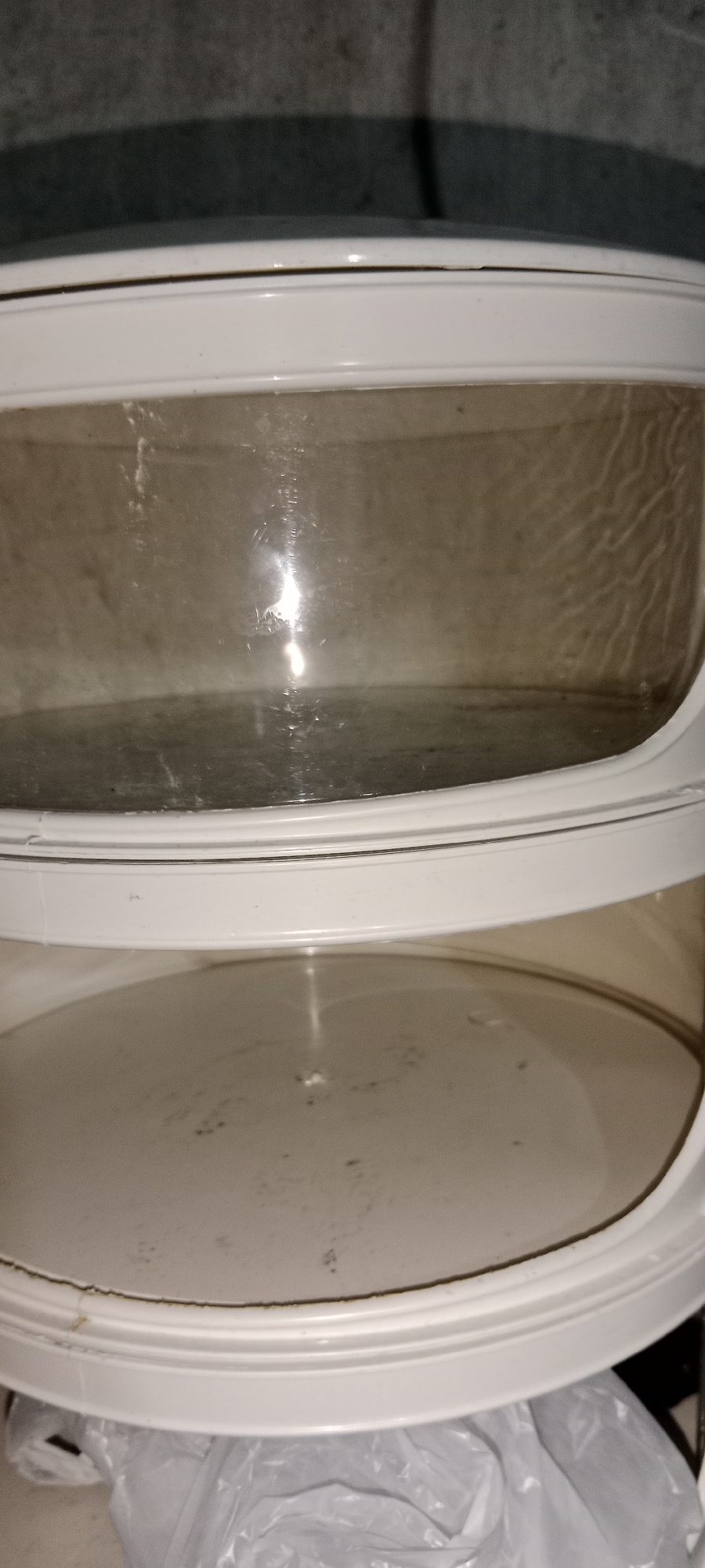 A two-tiered plastic food warmer is shown. The upper tier is a transparent container with visible dust and smudges, showing a reflective light source within. The lower tier is an opaque, off-white plastic base with some dark specks on its surface. The warmer is positioned on top of a crinkled white plastic bag, and the background is a dark, textured surface casting a shadow. The scene appears to be indoors, with no specific urban features of Subang, Indonesia visible.