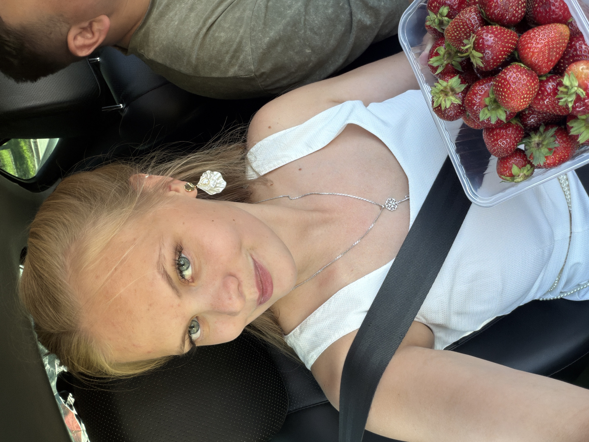 A young woman with blonde hair pulled back in a bun and blue eyes is taking a selfie while sitting in the passenger seat of a car. She is wearing a white top and a silver necklace with a key pendant. She is also wearing a large, shell-shaped earring in her left ear. In her right hand, she holds a clear plastic container filled with fresh strawberries. A man with short dark hair is visible in the background, sitting in the driver's seat. The car appears to be moving as greenery is visible through the window. The interior of the car shows black leather seats and a seatbelt across the woman's chest. There are no identifiable landmarks to place this image specifically within Izhevsk, Russia.