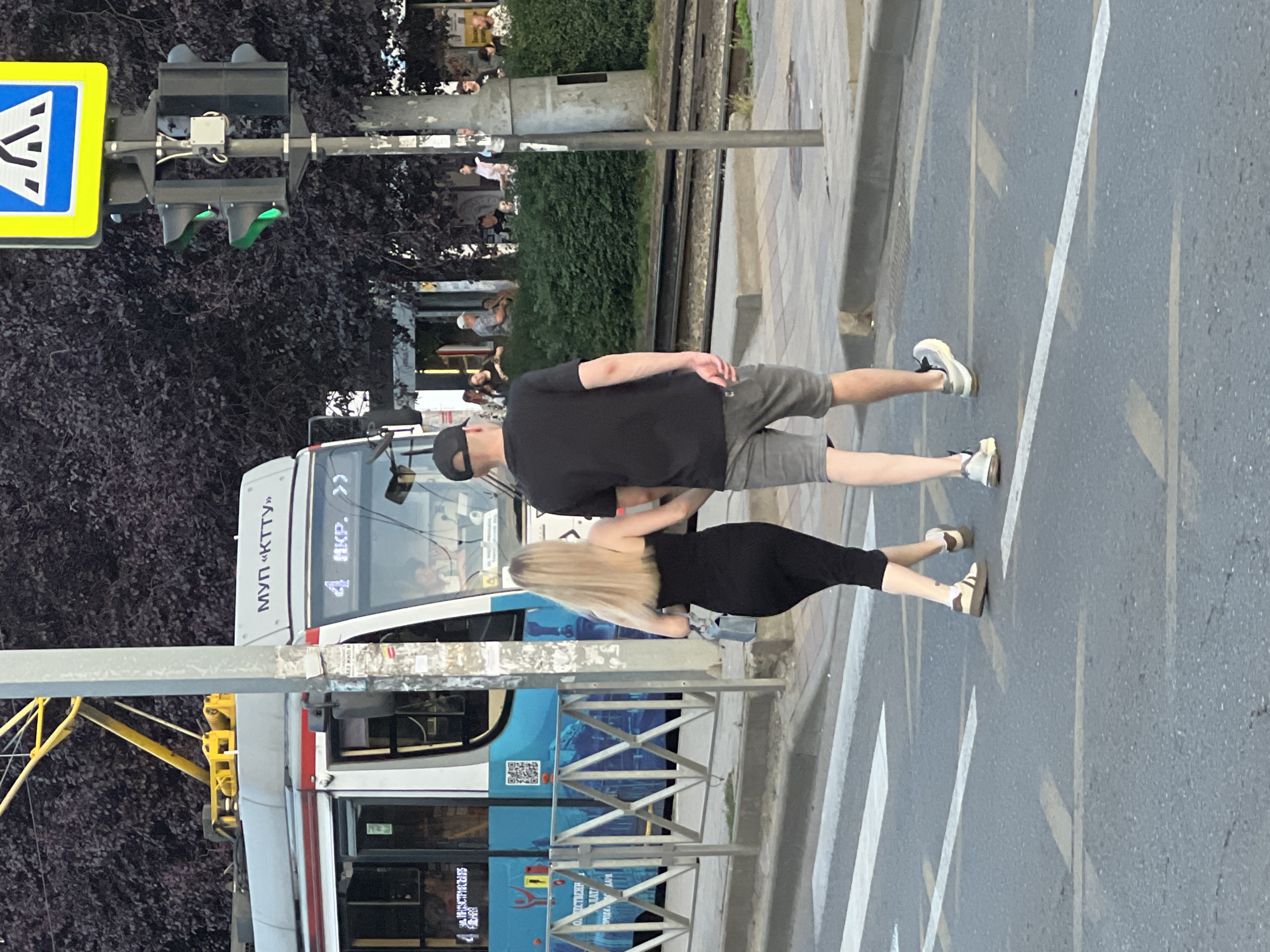 A young Caucasian couple, a man and a woman, are walking across a crosswalk in Krasnodar, Russia. The man, wearing a black t-shirt, black baseball cap, and grey shorts, walks with his left arm linked through the woman's right arm. The woman, with long blonde hair, wears a black strapless dress and carries a light blue purse. They are walking away from the viewer and towards the right side of the frame.
In the background, a modern tram with the route number "4" displayed on its front is stopped at a tram stop. The tram is white with blue and silver accents. The text "МУП «КТТУ»" is visible on the top front of the tram. To the right of the tram, a traffic light shows a green signal. Behind the tram and traffic light, a dense row of dark green trees and some buildings with people gathered outside can be seen. The ground is paved with asphalt, and the crosswalk lines are clearly visible. The overall scene appears to be a busy street in an urban environment.
