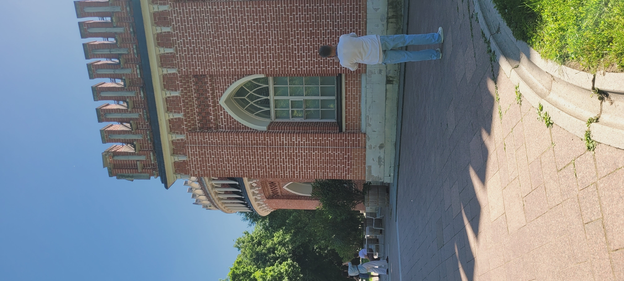 The scene depicts an outdoor area within the Tsaritsyno Museum-Reserve in Moscow, Russia, specifically featuring a Gothic Revival style building.
The building, constructed primarily of red brick with white mortar, occupies the central and right portions of the image. Its roofline is adorned with a crenellated parapet featuring regular merlons and a light-colored frieze below. A prominent pointed arch window, with white framing and intricate mullions, is visible on the main facade. To the left, a curved section of the building features an open arcade or colonnade with slender white columns and heart-shaped decorative elements above. The building rests on a light-colored stone foundation exhibiting signs of weathering and discoloration.
In the foreground on the right, a male individual stands facing away from the viewer, wearing a white t-shirt, light blue jeans, and white sneakers. He holds a device, possibly a camera or phone, directed towards the building. A blue strap is visible around his neck. In the mid-ground on the left, a female individual, dressed in a light blue top and light-colored wide-leg pants, stands holding a camera or phone up to her eye, appearing to photograph the building. Further in the background on the left, two other individuals are seated at outdoor tables, partially obscured by green foliage.
The ground is paved with light-brown rectangular pavers, displaying distinct, elongated shadows cast from the left. A curved concrete drainage channel delineates the edge of the paved area on the right, bordering a patch of green grass. Several small, potted or planted trees are arranged near the base of the building. Mature green trees are visible in the background on the far left. The sky is clear and bright blue, occupying the upper third of the frame, with a faint white speck visible in the upper left. The scene is brightly lit by direct sunlight.