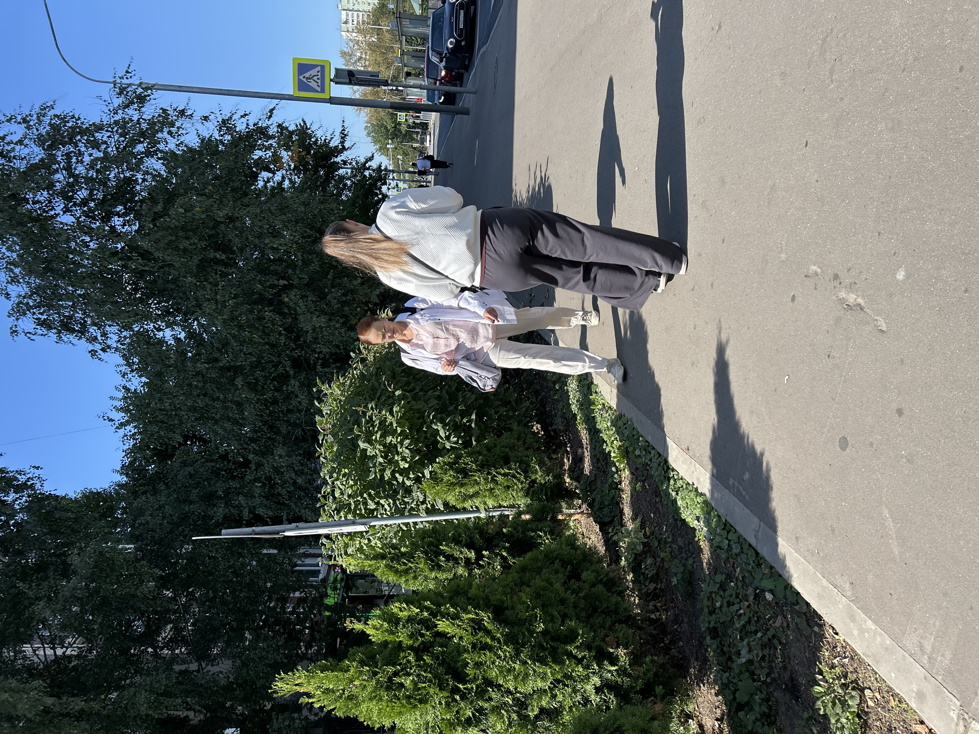 Two women are walking on a paved sidewalk next to a road in Moscow, Russia. The woman in front is wearing a light pink shirt and white pants, with glasses and red hair. She is carrying a grey bag over her shoulder. The woman behind her is wearing a white sweater and dark grey wide-leg pants. She has blonde hair pulled back in a ponytail and a black strap across her shoulder. The sidewalk is next to a strip of green vegetation including evergreen bushes and trees, and a larger tree with dark green leaves. A lamppost with a pedestrian crossing sign is visible behind the women. In the background, cars are parked along the side of the road, and buildings and trees are visible. The sky is blue and clear.