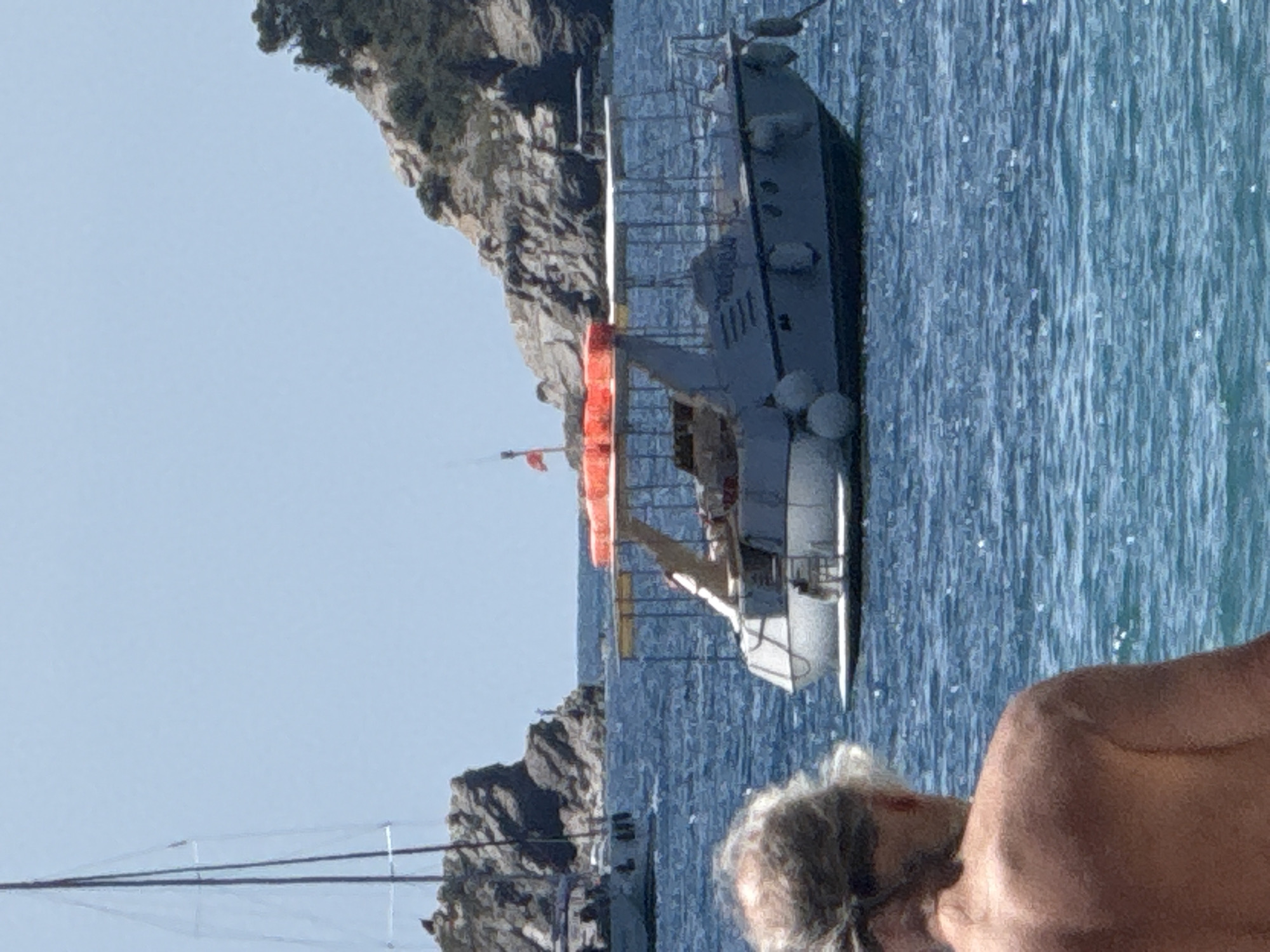 The image shows a sunny day with a clear blue sky over the sea. In the foreground, a person's back is visible, with their shoulders and head turned away from the camera. The person has light-colored hair tied back. In the mid-ground, a white boat with a red-orange canopy is on the water. The boat has a railing around its deck and several fenders along its side. The name "PIRATA" is visible on the side of the boat. Behind the boat, a rocky, tree-covered island rises from the sea. Another sailboat with a tall mast is visible in the background to the left, near a rocky outcropping. The overall scene depicts a coastal or island setting, likely a popular tourist destination, given the presence of recreational boats and clear weather. The image does not contain enough information to determine the specific location in Budva, Montenegro.