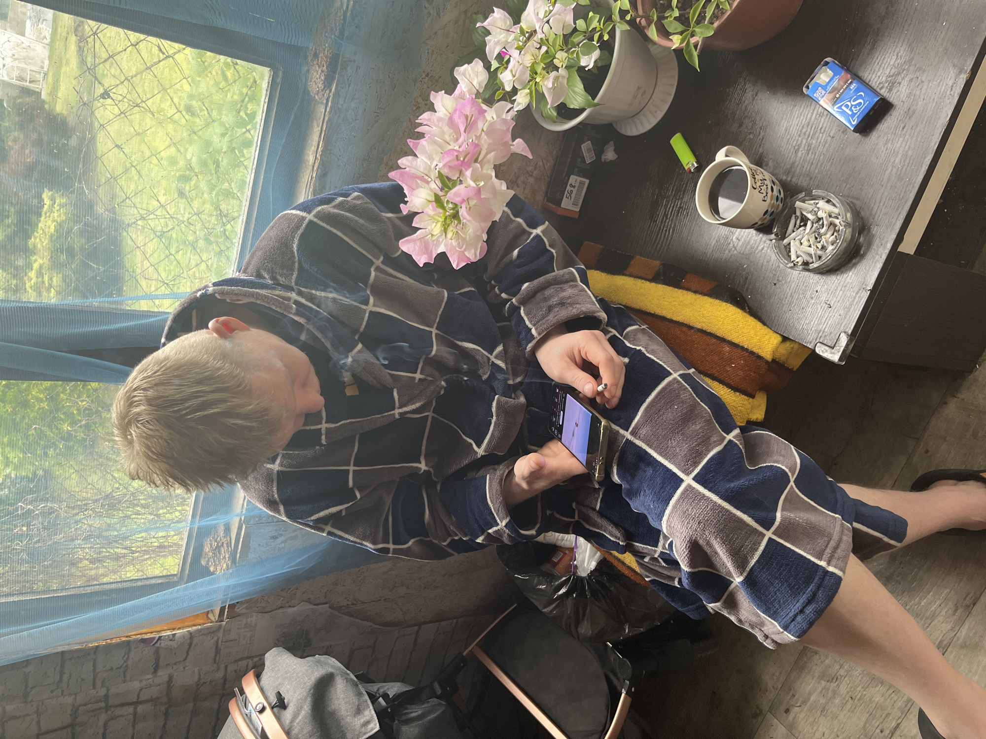 The image shows an individual in Vesyoloye, Russia, wearing a blue and gray checkered robe, sitting indoors near a window with a translucent blue curtain. The person is holding a smartphone in their left hand and a lit cigarette in their right. Next to them, on a dark-colored table, are potted flowers, a full ashtray, a cup of coffee, a lighter, and a pack of cigarettes. Below the individual, a gray bag and what appears to be a disassembled stroller frame are visible on the floor. The scene suggests a relaxed, indoor setting.