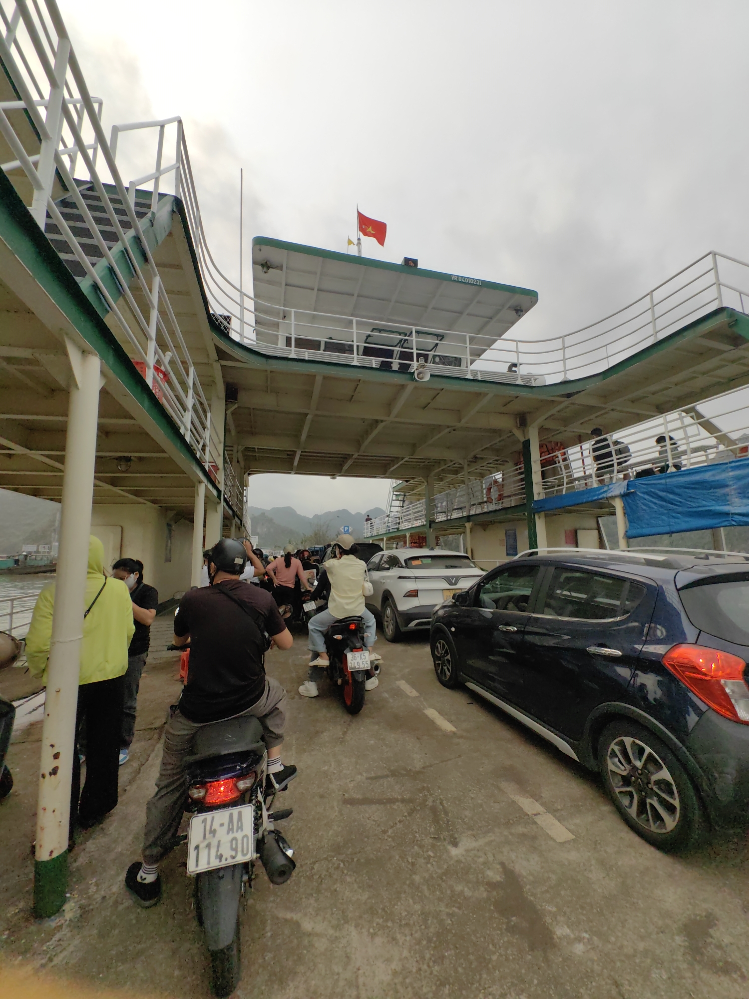 On a gray, overcast day in Hải Phòng, Vietnam, the scene depicts a crowded ferry. A queue of vehicles, including cars and motorbikes, is waiting to disembark. Two individuals on motorbikes, one with license plate "14-AA 114.90" and the other with "36-K5 429.55", are prominent in the foreground. To the left, a person in a yellow jacket and a face mask is visible. The ferry structure features white railings, green trim, and a red Vietnamese flag on top. Passengers are visible on the upper deck. The environment suggests a waterfront or river crossing location.